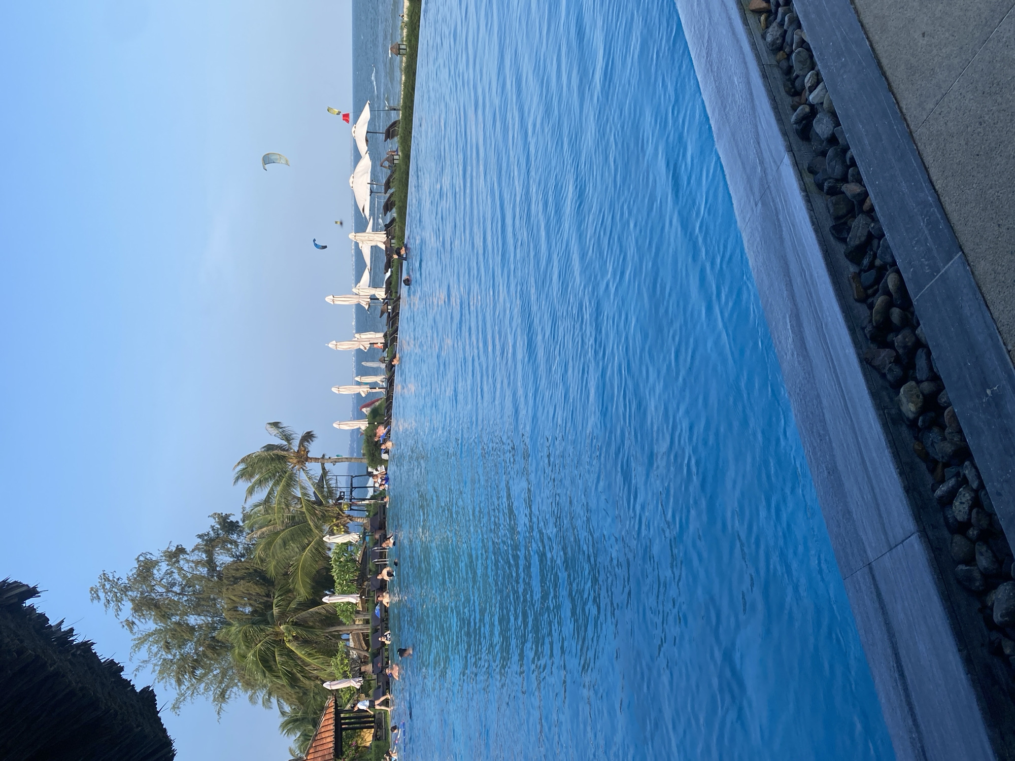 An outdoor scene in Phu Thuy Ward, Vietnam, depicts a resort environment under a clear light blue sky. In the foreground, a large, blue infinity pool occupies the lower two-thirds of the image, its surface showing ripples. The pool's edge is delineated by a dark grey stone coping, beneath which a trough holds rounded dark stones, adjacent to a concrete walkway. Multiple individuals are present in the pool, primarily at the far end, with some standing and others partially submerged.
Behind the pool, a strip of green foliage precedes a sandy beach area. This area is furnished with numerous white or cream-colored beach umbrellas, some open and some closed, alongside dark lounge chairs. Several people are visible on the beach, including one individual walking from left to right near a building with a reddish-brown tiled roof on the left side of the midground. Lush tropical vegetation, consisting of multiple palm trees and a taller, slender tree with fine green foliage, dominates the midground. A raised structure, resembling an observation platform, is also visible among the trees and umbrellas.
The background features the ocean with light waves breaking near the shore. Above the water, at least four kitesurfing kites are airborne; their colors include light blue, yellow-green, white/grey, and one with red accents. The sky is uniformly clear. A dark, thatched roof partially extends into the top-left corner of the image, indicating the vantage point.