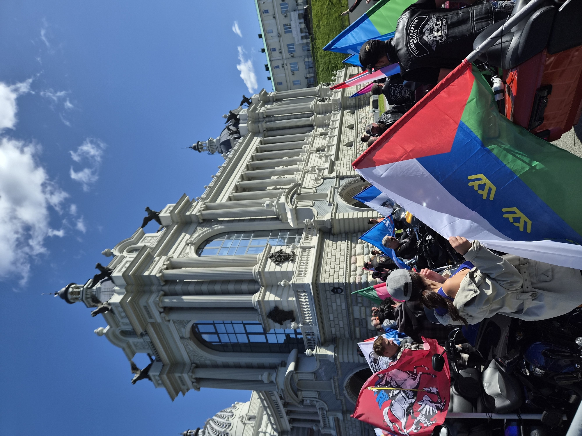 The image depicts a gathering of people in front of the Kazan Family Center (also known as the "Chalice") in Kazan, Russia. The foreground is dominated by several motorcycles and an array of flags, some of which are held aloft by individuals. Prominent among the flags are those with a red background featuring a white horseman, a tri-colored flag with blue, red, and green sections, and a blue flag. The people in the image are diverse in age and gender, with many wearing casual clothing, leather vests, and hats. They appear to be participants in an event, possibly a motorcycle rally or parade, given the presence of the motorcycles. The Kazan Family Center, a distinctive white building with classical architectural elements and a dome, serves as the backdrop. The sky is clear and blue, with a few scattered white clouds. The overall scene suggests a sunny day and a public gathering.