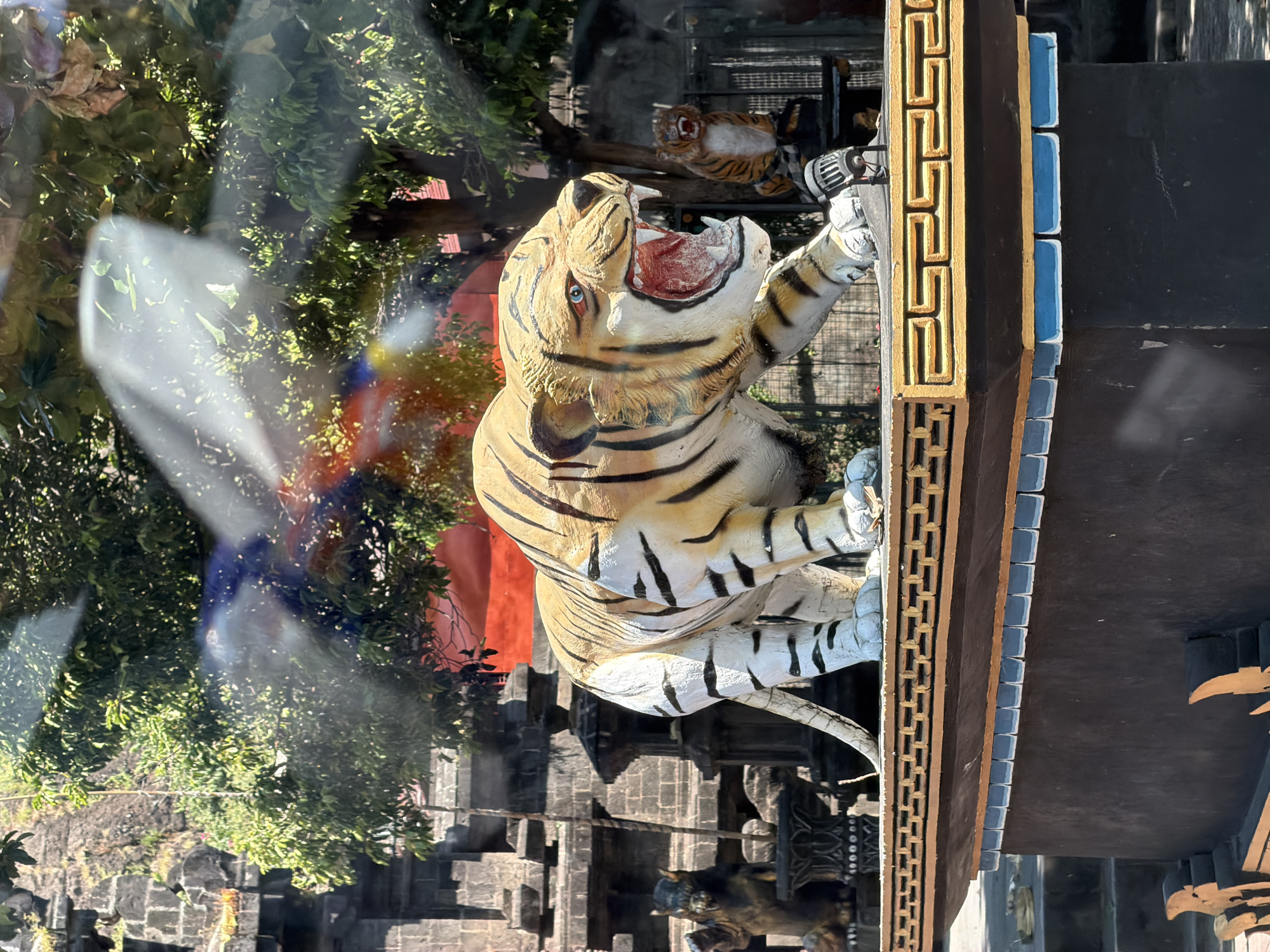 The image shows a vertical view of a tiger statue clinging to a building. The tiger is cream-colored with black stripes, baring its teeth, and with its paws against the wall. A second, smaller tiger statue is visible behind the larger one. The building is a combination of black, gold, and blue. The surrounding environment includes a structure with gray stones, likely part of the temple complex. Green trees are visible in the background, with a reflection. The setting is likely a temple located in Banyu Poh, Indonesia.