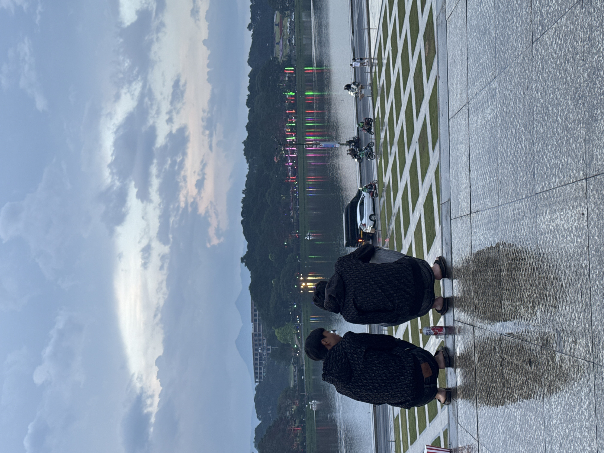 The image shows a lakeside scene in Da Lat, Vietnam. Two individuals are crouched near the water's edge, their backs to the viewer, wearing dark patterned jackets and sandals. In the background, across the lake, are colored lights and reflections in the water, possibly indicating a festive or illuminated area. Parked motorbikes and a white car are also visible. The immediate foreground consists of a tiled surface with a grid pattern of grass inserts. The sky is cloudy with hints of sunset colors.