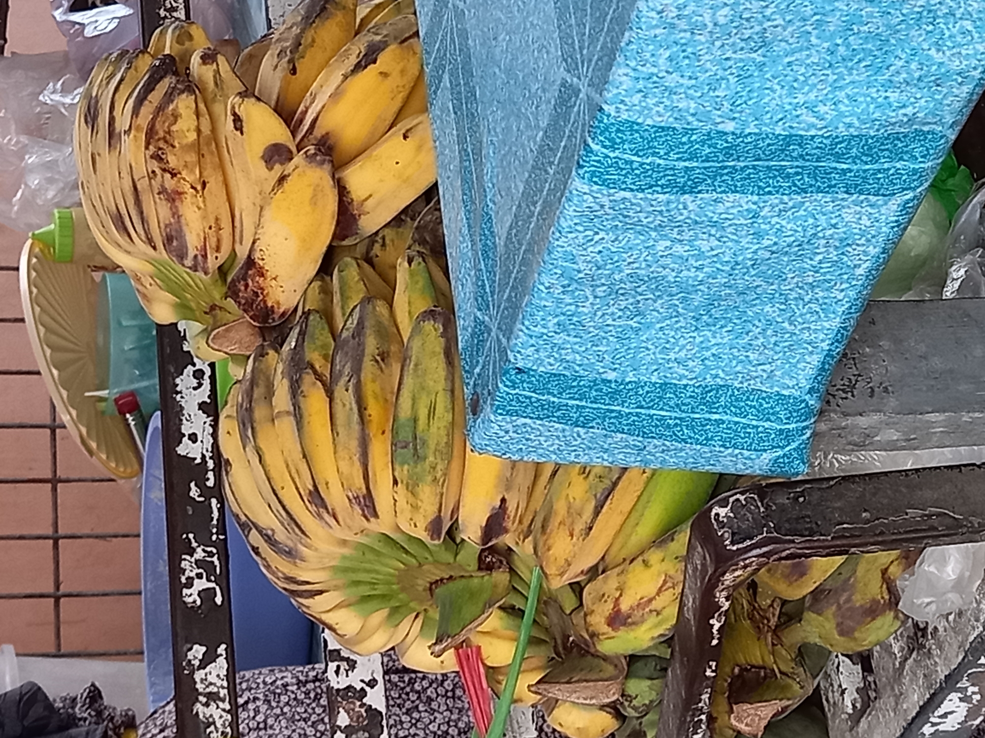 A close-up shot captures a bunch of ripe bananas, some with brown spots, piled on a metallic surface. A blue cloth with stripes covers a portion of the scene. The background reveals a tiled wall and a glimpse of a basket. The image is believed to be taken in Samarinda, Indonesia, likely at a market given the presence of produce and market-like structures.