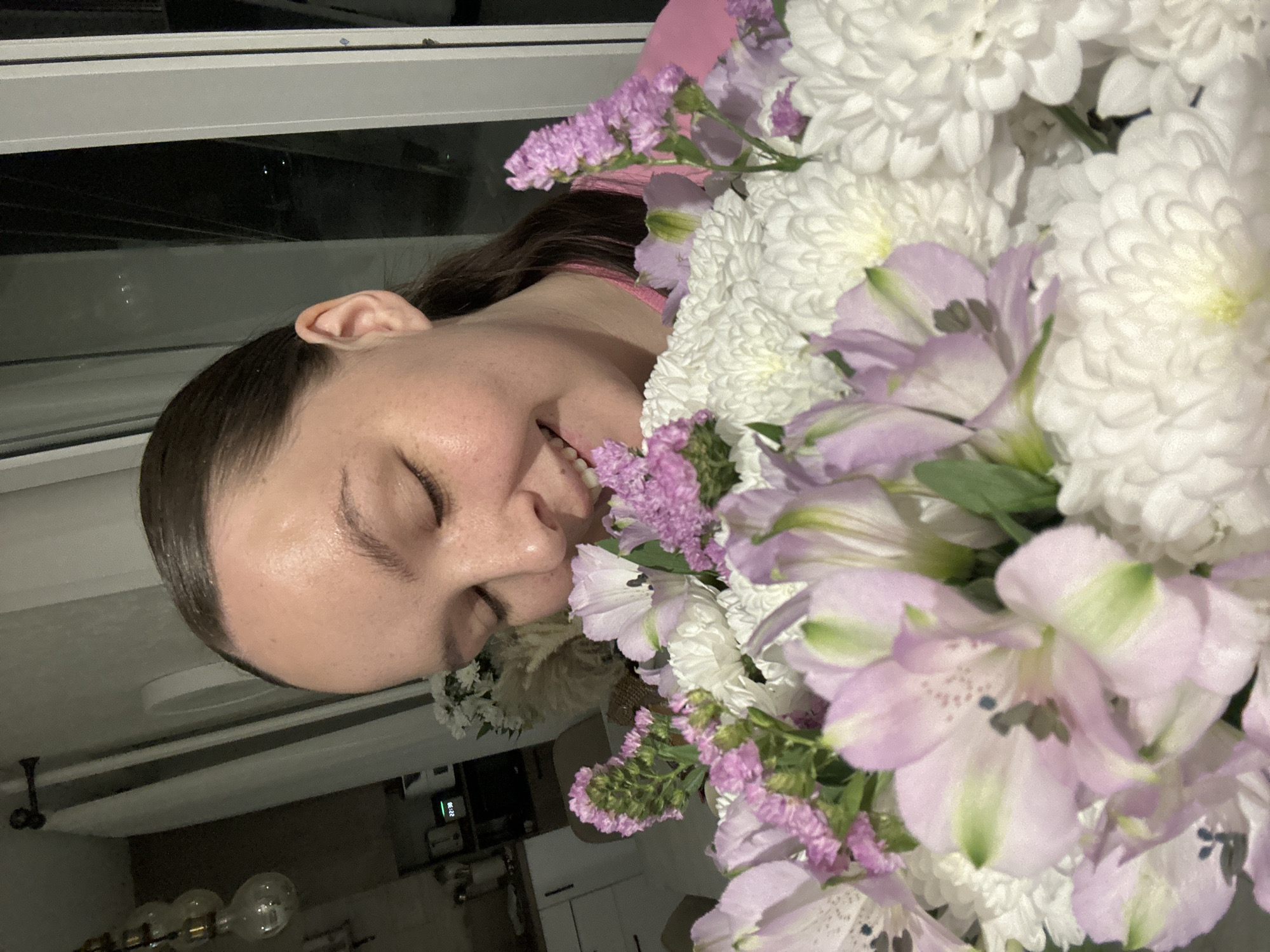 A woman with brown hair smiles with her eyes closed amidst a large bouquet of flowers, seemingly indoors. The flowers include white chrysanthemums and light purple alstroemeria. Visible in the background through a window are other structures, possibly buildings, and a lit-up room with a hanging light fixture. A digital clock in the lower part of the image reflects a time of 22:30. The location is unconfirmed to be in Chaikovsky, Russia.