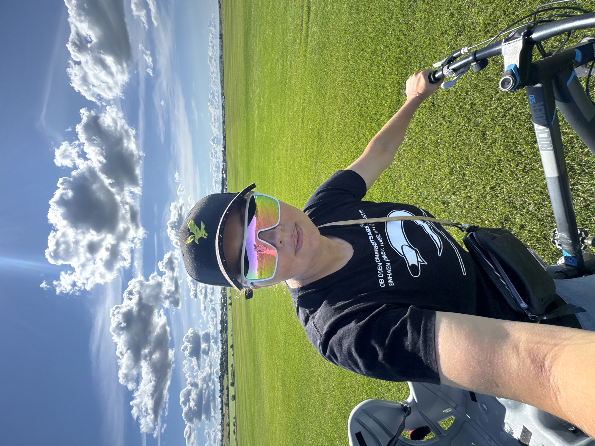 A medium shot photo shows a woman with fair skin and brown hair, wearing a black t-shirt with a graphic print, a black baseball cap with an embroidered golden eagle, and large white sunglasses with a rainbow lens, seated on a bicycle. The woman is positioned slightly off-center to the right, facing the camera and holding the bicycle's handlebars. Her left arm is bent, resting on the handlebars, while her right arm is extended. A gray child's bicycle seat is visible on the rear of the bicycle, to the left of the woman. She is also wearing a black cross-body bag with a zippered compartment. The background is a vast, bright green grassy field under a bright blue sky with scattered white clouds. In the distance, a line of trees and small buildings can be seen along the horizon. The image appears to have been taken outdoors in Kempelevo, Russia, on a sunny day.