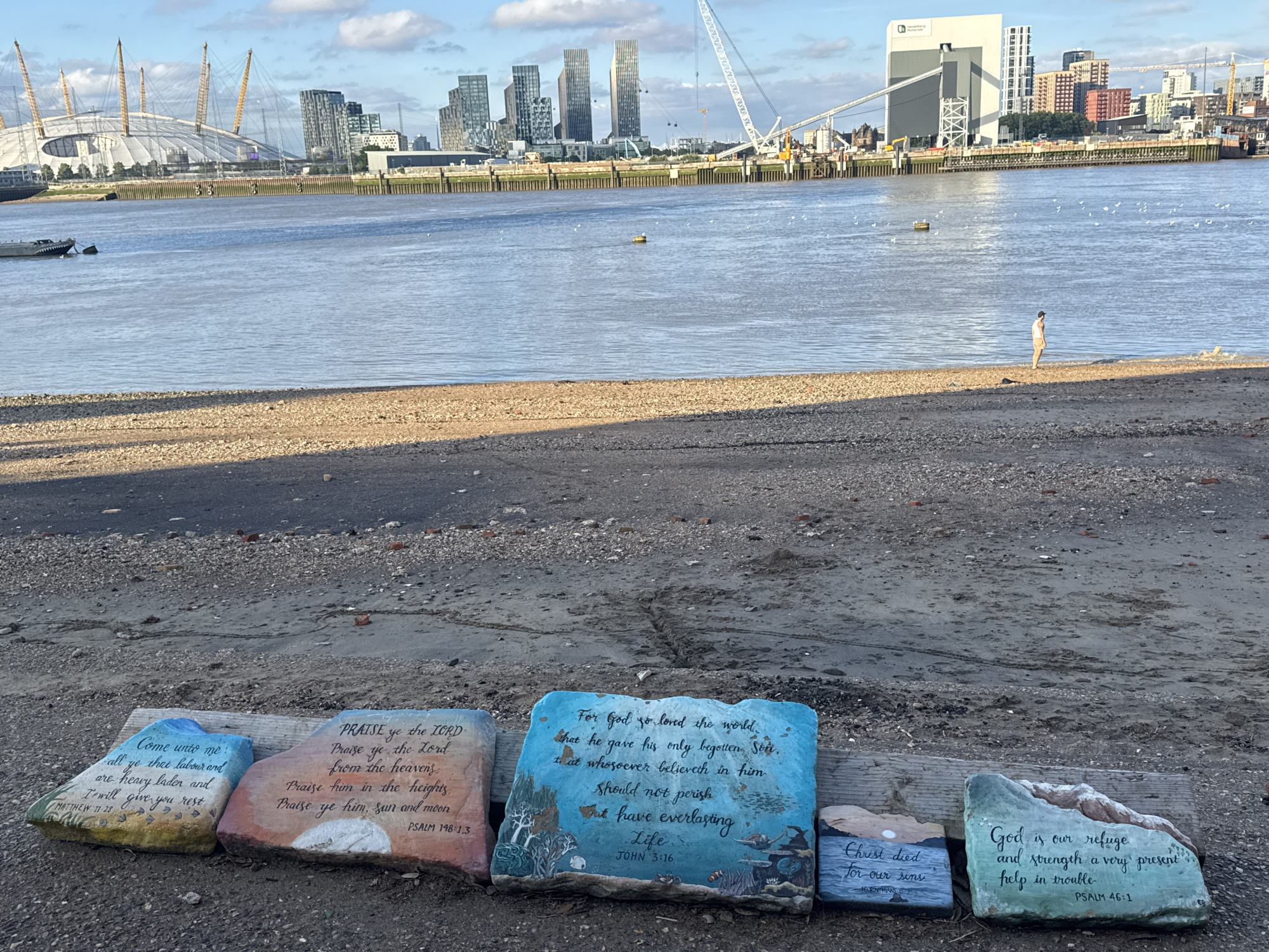 The image depicts a section of the River Thames waterfront in London, United Kingdom. In the foreground, several large, flat rocks are arranged on a sandy and gravelly shore, each painted with religious verses and illustrations. One rock features a blue background with imagery of marine life and trees, and text that reads, "For God so loved the world, that he gave his only begotten Son, that whosoever believeth in him should not perish, but have everlasting life. John 3:16." Another rock, with a pink and orange sunset background, has text stating, "Praise ye the Lord from the heavens, Praise ye him, sun and moon. PSALM 148:1,3." Adjacent to this is a rock with a blue and white gradient, containing the text, "Come unto me, all ye that labour and are heavy laden and I will give you rest. MATTHEW 11:28." Further to the right, a greenish rock has a verse that says, "God is our refuge and strength a very present help in trouble. PSALM 46:1." A smaller rock to its left reads, "Christ died for our sins. 1 CORINTHIANS 15:3."
In the midground, the calm waters of the River Thames are visible, with several small boats moored. On the opposite bank, the London skyline is prominent, featuring distinctive modern architecture. Notable landmarks include the O2 Arena with its iconic dome structure and multiple yellow masts, and a cluster of tall residential or office buildings. A large white construction crane stands prominently against the sky, suggesting ongoing development or construction in the area. The sky is partly cloudy, with the sun casting light on the scene. A solitary figure, appearing to be a person standing on the shore, is visible in the distance, looking out towards the river. The overall environment is a blend of urban landscape and natural elements, with the waterfront serving as a focal point.