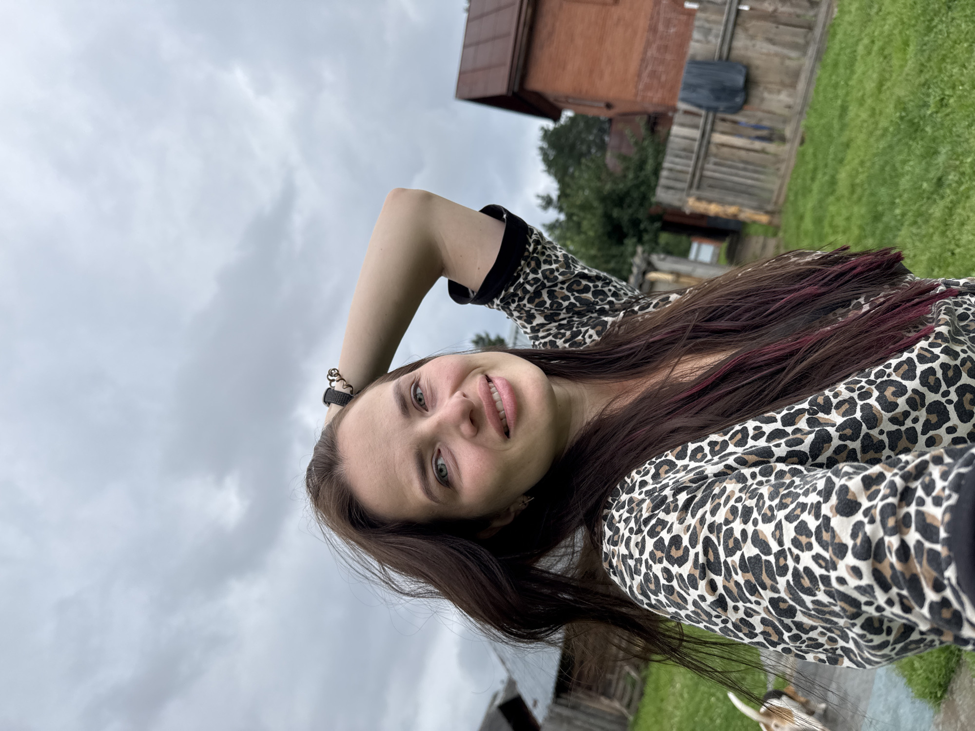 A medium shot from a slightly low angle captures a woman in a leopard-print top standing outdoors. Her left arm is raised, bent at the elbow, with her hand resting on the back of her head. She has long, dark brown hair with some pink highlights. The background consists of a cloudy sky, green grass, wooden fences, and rustic buildings, suggesting a rural or suburban setting. A small dog, primarily white with brown markings, is visible on the left side of the frame, facing away from the camera.