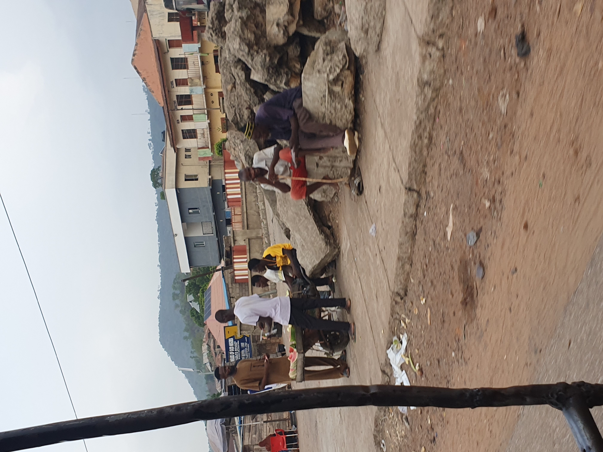 This image captures a daytime street scene in Lokoja, Nigeria, under an overcast sky. The overall setting appears to be a bustling urban or semi-urban area with hilly terrain in the background.
In the mid-ground, a group of approximately five to six men are gathered along a dirt and concrete pathway. On the right, two men are sitting among large rocks: one in a dark blue shirt and striped yellow cap, and another in a white shirt and red shorts holding a stick. Further left, a man in a white t-shirt stands next to a small, wheeled vendor cart, which holds sliced watermelon, suggesting he is a seller. Another man, dressed in a brown traditional outfit, stands nearby. Another man in a yellow shirt is also visible in the group. The ground is somewhat unkempt with scattered litter, including paper and plastic.
The background features several multi-story buildings of varying colors, including beige, light blue, and red-roofed structures, some with balconies. Behind these buildings, hazy green hills rise into the distance. Power lines stretch across the upper part of the scene.
Notable details include the overcast weather, which softens the light, and the presence of vendor activity. Visible text on a sign reads "UNITED OF GOD NIGERIA" and "LOKOJA HQ," confirming the location. Another partial sign appears to say "SCHOOL." A dark, metallic railing or bar partially obstructs the very bottom foreground of the image.