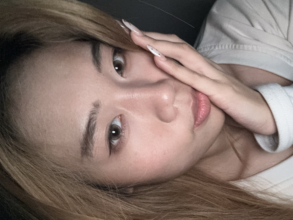 A close-up portrait captures a young East Asian female in a dimly lit indoor setting in Yuzhong District, China. The subject has fair skin and light brown hair parted on the left, framing her face. She wears a light-colored, long-sleeved garment. Her brown eyes appear enlarged due to cosmetic contact lenses and are accentuated with black winged eyeliner. Her eyebrows are well-defined. Her lips are slightly parted. Her right hand, adorned with long, sculpted, light-colored fingernails featuring small dark decorative dots, rests against her right cheek and jawline. She gazes directly at the camera with a neutral expression. The background is dark and indistinct.