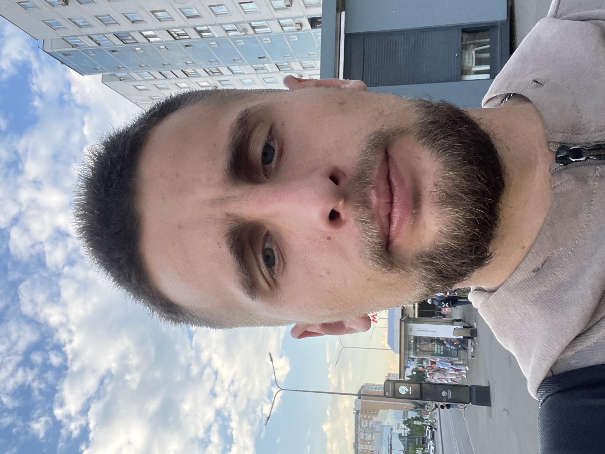 A close-up selfie of a man with short dark hair, a beard, and a light-colored hooded jacket is taken outdoors. The man is looking directly at the camera. In the background, there is a street with a bus stop, a tall building with blue and white facade, and a cloudy sky. The man is standing on a street with a sidewalk and a road with cars. A sign with a metro logo is visible at the bus stop. The overall scene suggests an urban environment in Moscow, Russia.