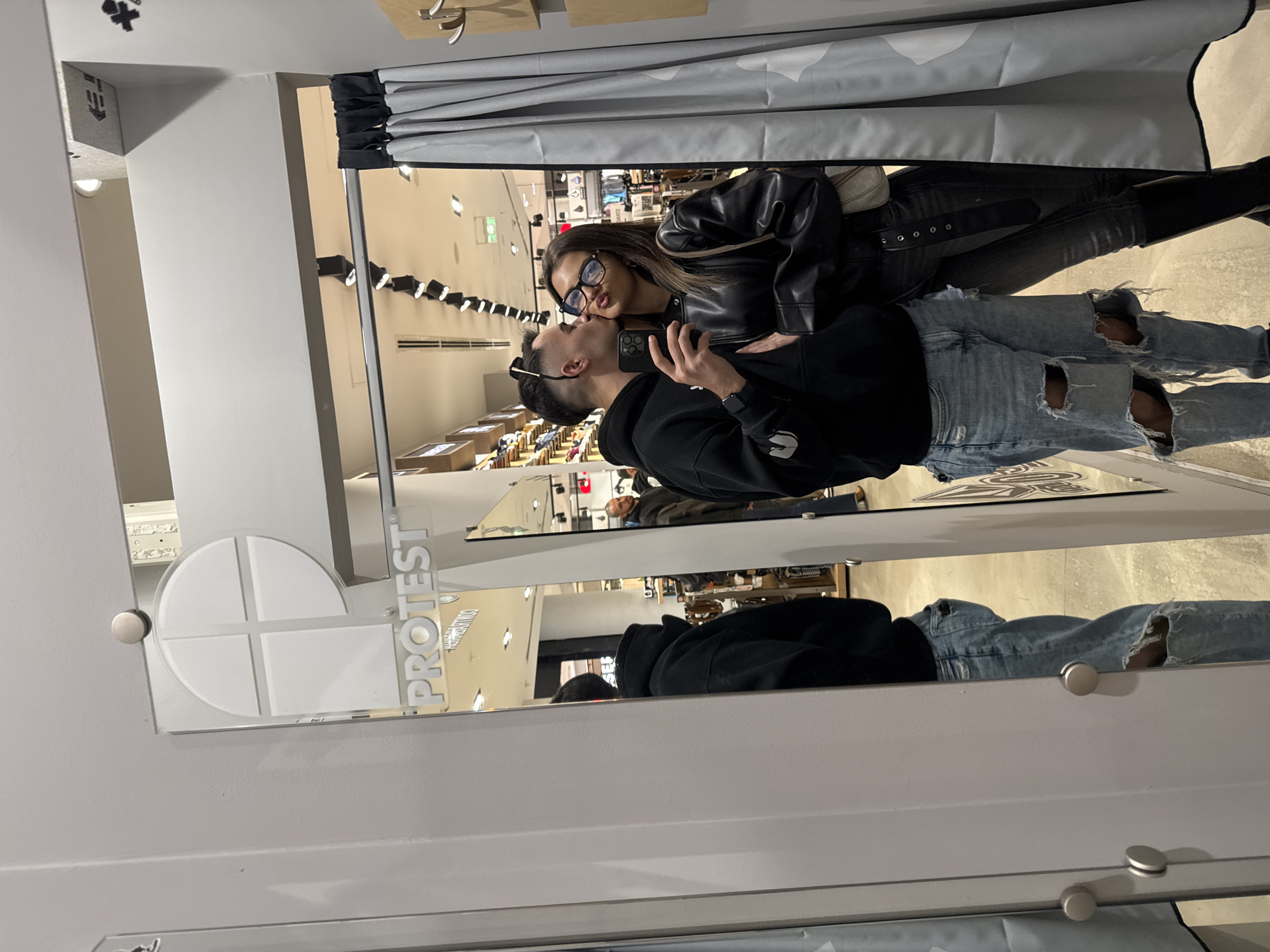 A photo, taken with a smartphone, captures a couple posing for a selfie in a retail environment, possibly in Sofia, Bulgaria. The pair stands within a fitting room framed by a white-painted structure with "PROTEST" inscribed on it. The woman wears glasses and a black leather jacket. The man is in a black hoodie, distressed jeans, and sunglasses perched on his head. They are kissing as they hold a phone for the photo. The background shows a store interior with retail displays and lighting fixtures. Clothing and fitting room curtains are visible above and behind the couple.