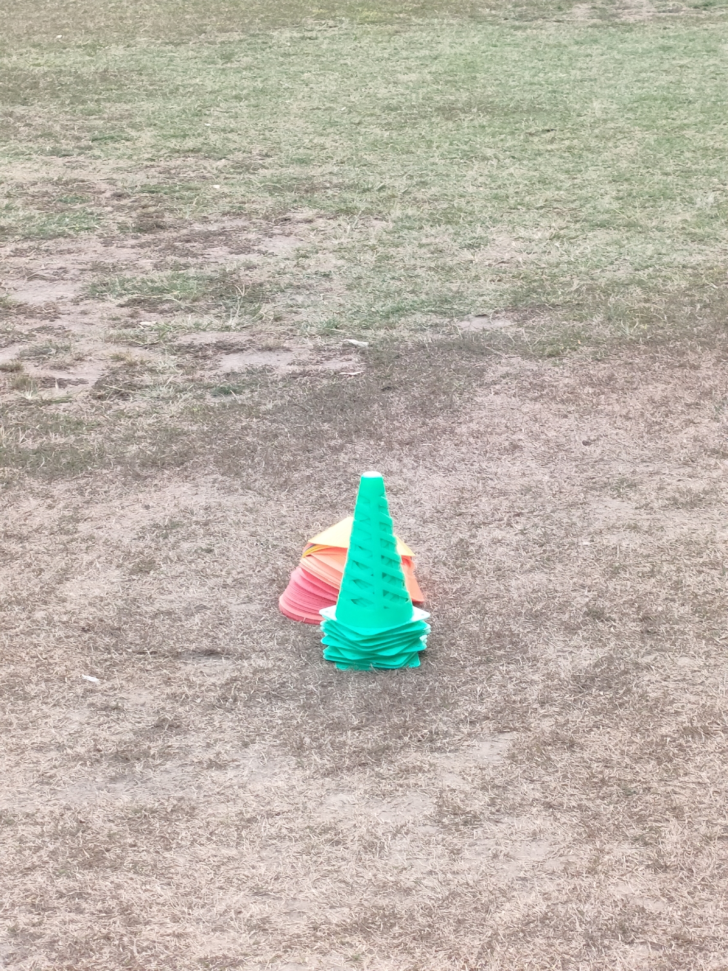A pile of training cones sits on a patch of dry grass in Banguntapan, Indonesia. The cones are stacked, with green cones on top and orange and pink cones underneath. The grass is sparse and dry, indicating a lack of recent rainfall. The area appears to be an outdoor training or sports field.