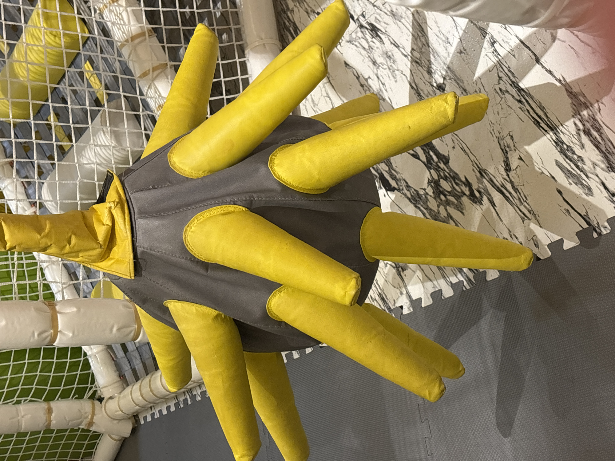 The image shows a stuffed toy with a gray body and yellow protrusions, resembling a spiky ball or a starburst. It is located indoors on a gray padded floor with a marble-patterned surface. In the background, a white net enclosure with padded white poles is visible. This appears to be part of a children's play area, likely in a soft play center. There is no indication of the city of Dubai, United Arab Emirates, or any specific location within it. The scene is static, with no people or actions depicted.