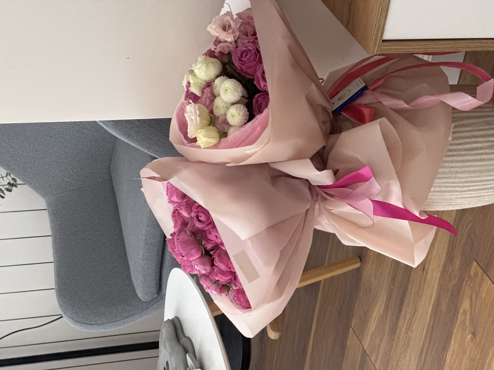 Two bouquets of flowers wrapped in pink paper and tied with pink ribbon sit indoors, likely in a home or office setting. One bouquet consists of pink roses, and the other contains a mix of pink and white flowers. The bouquets are placed on a white surface, with a gray chair visible in the background. The floor is made of wood. The location is not specified.