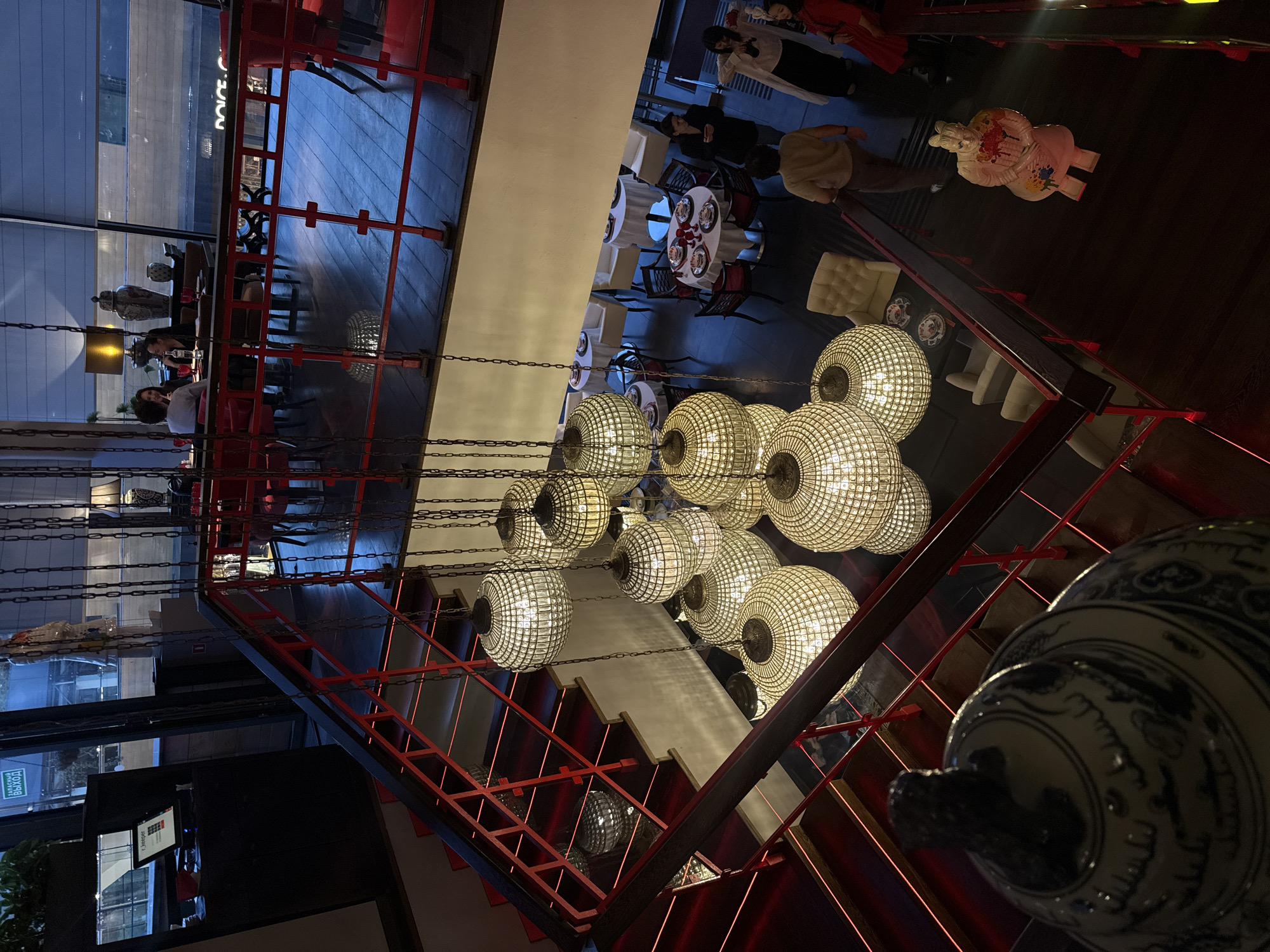 The image depicts the interior of a restaurant, possibly located in Barvikha, Russia, given the signage that appears to be in Russian. The scene is captured from an elevated perspective, looking down a wide staircase with a red railing. The staircase is illuminated with red LED strip lights along the risers.
Dominating the foreground and midground are several large, ornate pendant lights with a spherical, lattice-like design, casting a warm glow. The restaurant features dark wooden floors and a modern aesthetic with Asian-inspired decorative elements.
In the upper left, a reception desk with a computer monitor and signage is visible. Further back, through large windows, streetlights and building facades suggest an urban setting, possibly at dusk or night.
On a landing above, several tables are set for dining, with white tablecloths and red chairs. Some patrons are seated at these tables. In the midground, a group of people are gathered near a staircase. A person in a light brown shirt walks away from the camera, while two women converse. Another woman in a white coat and a person in a red dress are also present.
Near the bottom right of the image, a large, decorative statue resembling a Terracotta Warrior figure is positioned on the staircase. A large ceramic vase with blue and white intricate patterns sits in the lower left corner, partially obscuring the view of the staircase. The overall atmosphere appears to be dimly lit, with ambient lighting highlighting the decorative features and creating a sophisticated ambiance.