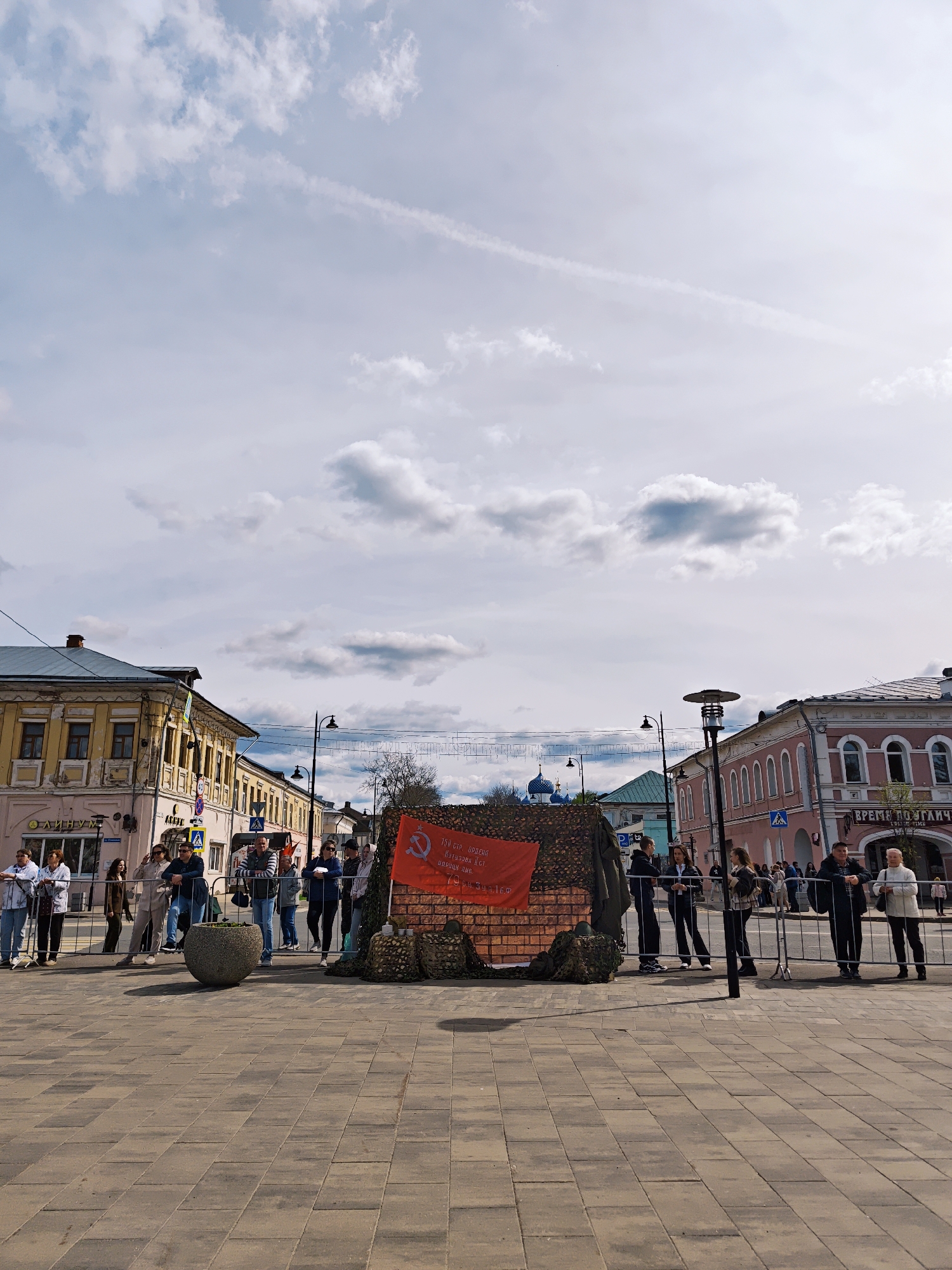 The image shows a staged historical exhibit in the city of Uglich, Russia. The exhibit appears to be a small red brick structure decorated with camouflage netting and sandbags. A red banner with a hammer and sickle symbol hangs on the structure. Text on the banner reads "150th Order of Kutuzov ISTR. IPRIZ DIV 70 S.V. Z U.A. 16.02". A metal fence is positioned in front of the exhibit, separating it from the pedestrian area. A group of people, including men and women of varying ages, are standing behind the fence, some leaning on it as they observe the display. The background features a paved square surrounded by multi-story buildings with architectural details. The sky is partly cloudy, with visible contrails from aircraft. Street lamps are placed along the square. Two commercial signs visible in the background, one reading "АлинУМ" and "ВРЕМЯ ПО УГЛИЧ" suggesting this location is a central square.