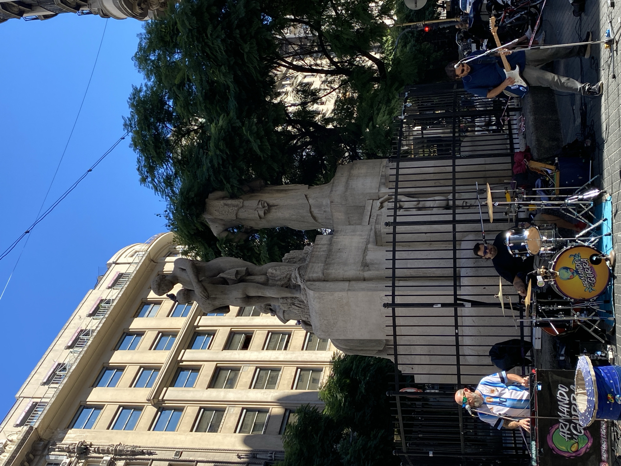 An outdoor music performance takes place in Buenos Aires, Argentina, under a clear blue sky. The scene is set on a paved street beside a tall, light-colored building with multiple windows, some of which appear to have external fire escapes or decorative elements. In the foreground, a multi-figure stone monument or statue, possibly depicting a group of people, stands behind a black metal fence. An inscription "PARA LA HUMANIDAD" (FOR HUMANITY) is visible on its base.
Three musicians are performing. To the left, a guitarist in a blue shirt, gray pants, and sunglasses plays a light-colored electric guitar. In the center, a drummer wearing a black shirt and sunglasses sits behind a drum kit. His yellow bass drum prominently features a logo with an alien head and the text "ARMANDO LA GORRA." To the right, a DJ or vocalist, identifiable by his beard, sunglasses, and an Argentina national football team jersey (blue and white stripes), stands behind a table with electronic equipment and a microphone. A blue hat is placed on his table, also bearing the "ARMANDO" name. A banner on his table reiterates the "ARMANDO LA GORRA" name and alien logo, along with a social media handle "@ARMANDOLAGORRA."
The bright sunlight suggests it is daytime, casting clear shadows. Lush green trees are visible around and behind the statue, adding to the urban park-like setting. Overhead wires crisscross the blue sky. The overall impression is a lively street performance, possibly busking, given the name "Armando la Gorra," which translates to "Armando the Cap" and is commonly associated with collecting donations for street art.