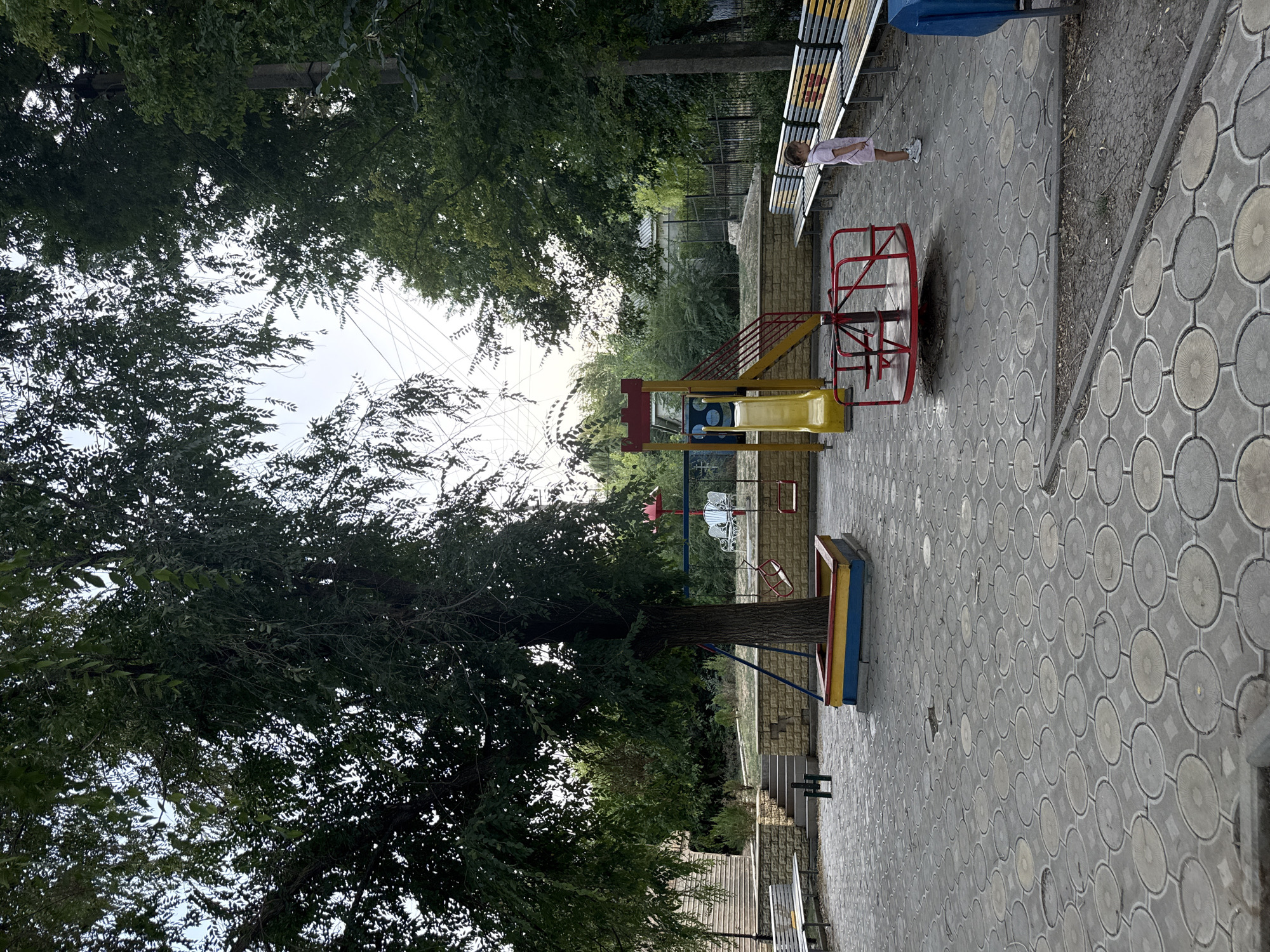 This is a full-shot, eye-level, outdoor photograph taken during the day in a public park or playground in Basarabeasca, Moldova. The scene is dominated by a large tree in the center, with lush green foliage. The ground is paved with circular concrete tiles, and a portion of the circular pattern is visible in the foreground.
To the left of the tree, a white bench is positioned near a low, stone wall with steps leading up behind it. To the right of the tree, a red and yellow swing set is visible. Further to the right, a yellow playground slide with red railings is set against a stone wall. In front of the slide, a red merry-go-round is present. A young girl, wearing a light pink outfit, stands near a row of yellow and black striped benches on the right side of the frame, holding a stick. A blue garbage can is partially visible on the far right edge of the image. In the background, more trees, a fence, and buildings can be seen. The sky above is a pale blue with some visible electrical wires.