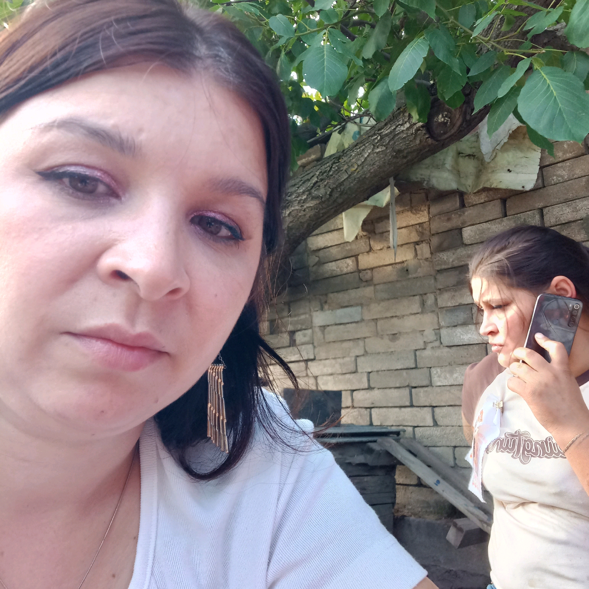 The image, captured in the city of Klyntsi, Ukraine, depicts two women outdoors. The woman in the foreground is looking directly at the camera, her face partially visible from the nose up. She has dark hair and is wearing makeup, including pink eyeshadow and eyeliner. She is also wearing a white top and a gold-colored earring. In the background, the second woman is holding a smartphone to her ear and appears to be engaged in a phone conversation. She is wearing a light-colored t-shirt with a graphic print. The setting includes a brick wall, a tree with green leaves, and some wooden structures, suggesting an outdoor, possibly residential or yard area. The lighting indicates it is daytime.
