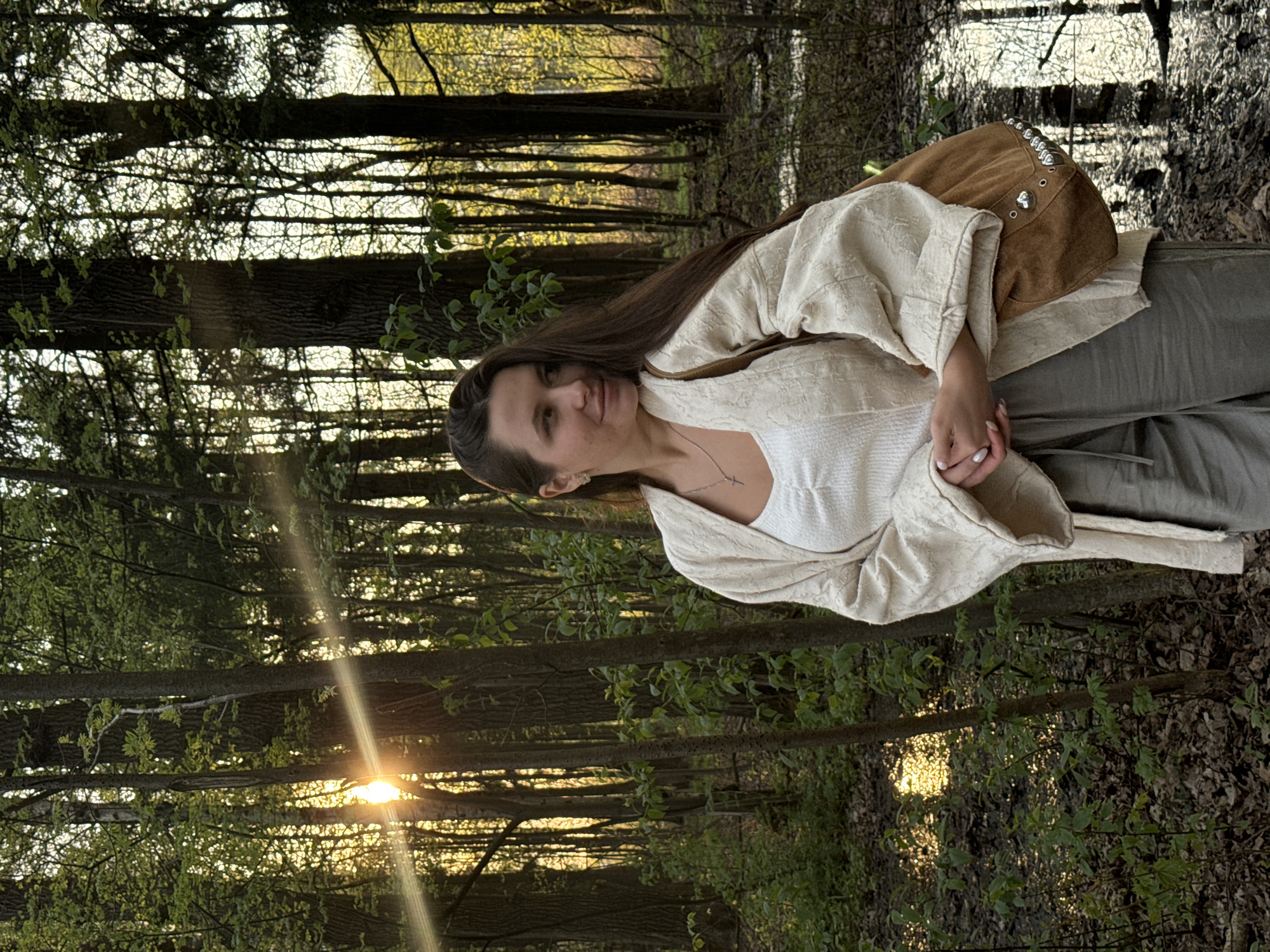 A woman stands in a wooded area, possibly in Khimki, Russia. She has long brown hair and is wearing a white top, a cream-colored robe, and gray pants. She has a brown suede bag with silver embellishments over her shoulder. Her hands are clasped together. The sun is shining through the trees in the background. The ground is covered in leaves and there is a body of water in the distance.