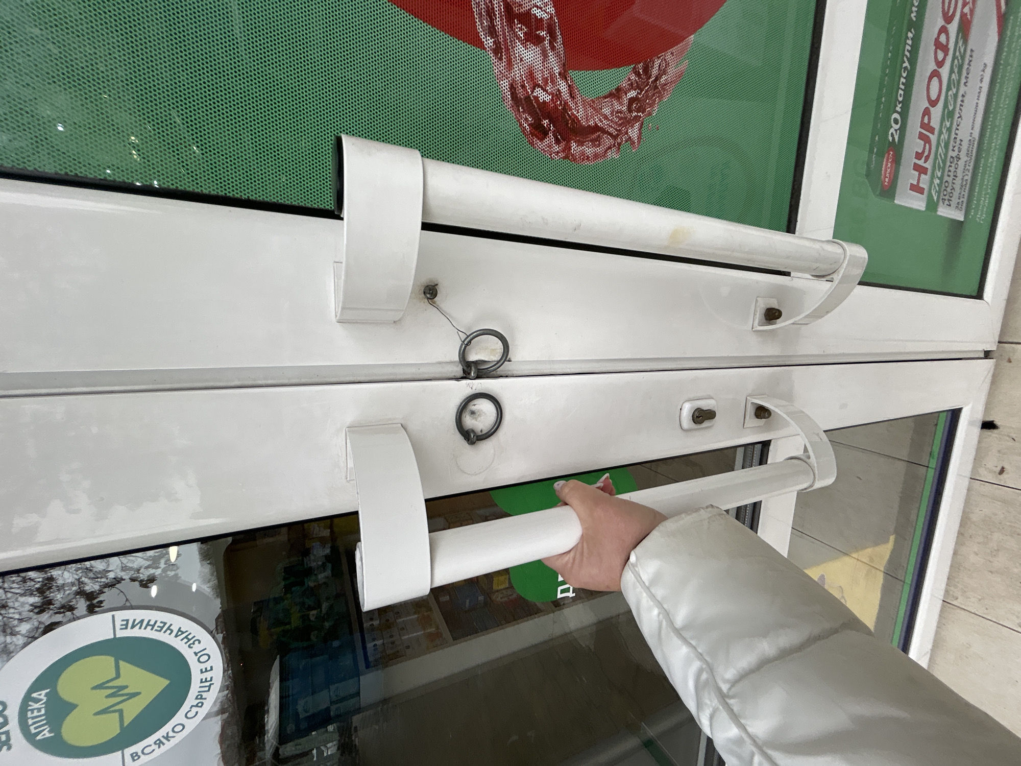 A person is opening the white door of a pharmacy. The door is made of glass and has white handles. The pharmacy has green and red advertisements on the window. A sticker with "Apteka" (Pharmacy) and "Vsjako syrtse e ot znachenie" (Every heart matters) in Bulgarian is on the window. An advertisement for "Nurofen Express Forte" in Bulgarian is also visible on the window. The person is wearing a white jacket. It is daytime. The location may be Plovdiv, Bulgaria.