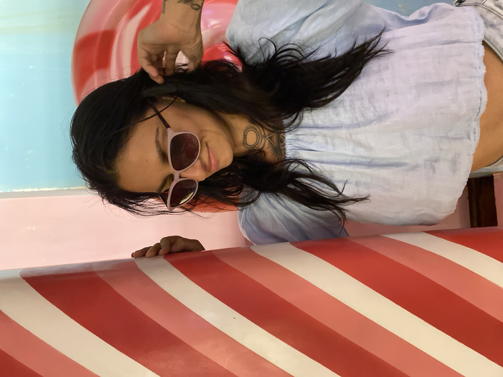 A medium shot captures a young woman wearing light blue off-the-shoulder top and sunglasses, standing against a colorful background. She is partially obscured by a large, red and white striped cylindrical object on her left. Her right hand is raised to her dark hair. A tattoo is visible on her right arm and neck. The background features soft pink and light blue hues, with a hint of a large red and white circular object. The scene is indoors, likely in a playful or decorative setting, and has been identified as being in Tien Thanh Ward, Vietnam.