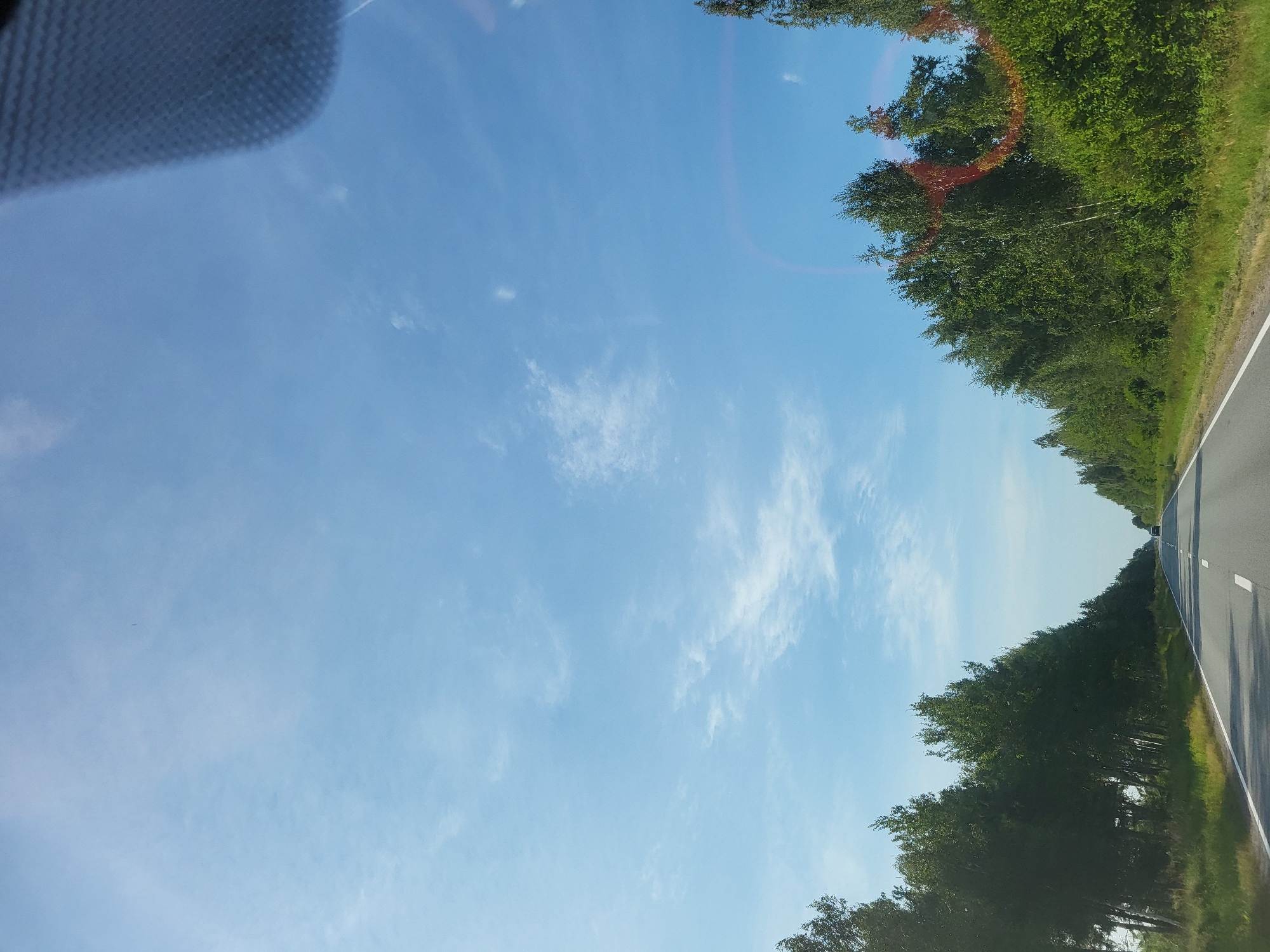 The image depicts a road passing through a rural landscape with trees lining both sides. The sky is a clear blue with scattered white clouds. A vehicle is visible in the distance on the road. The scene appears to be a daytime shot, likely taken from a moving vehicle, as evidenced by the slight blur and the perspective. The location is identified as Poķi, Latvia. The overall impression is one of a peaceful drive through a natural setting.