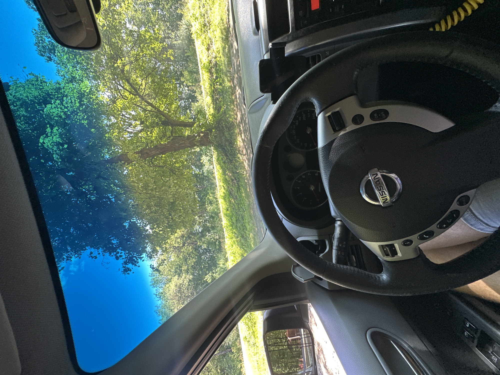 The image shows the interior of a Nissan vehicle, looking forward through the windshield. The view outside is of a tree-lined area with a road or path and green grass. Sunlight is filtering through the leaves of the trees. Inside the car, the steering wheel is visible, with the Nissan logo in the center. Parts of the dashboard and door panel are also in view. There are no people clearly visible in the image, but legs of a person are visible in the driver's seat. The specific location in Shatava, Ukraine, cannot be determined from the image itself.