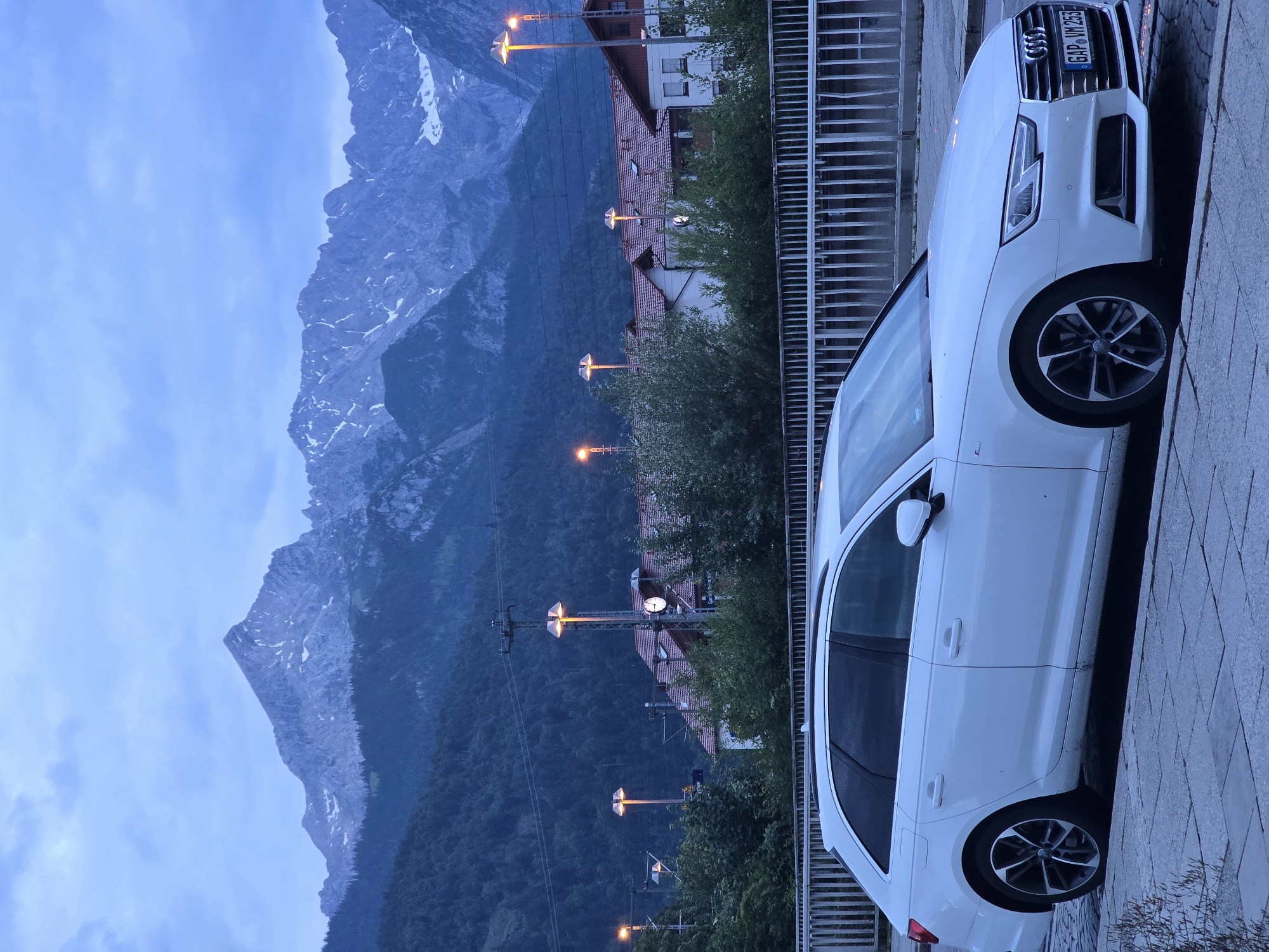 The image depicts a white Audi station wagon parked on a cobblestone street in Garmisch-Partenkirchen, Germany. In the background, a dramatic mountain range with snow-capped peaks dominates the skyline under a cloudy, twilight sky. Several lampposts with warm lighting are visible, illuminating a row of buildings with red-tiled roofs, suggesting a train station or a similar public facility. Dense green forest covers the lower slopes of the mountains. The license plate of the car is visible, reading "GAP VM 253". The scene is serene and captures the essence of a European alpine town at dusk.