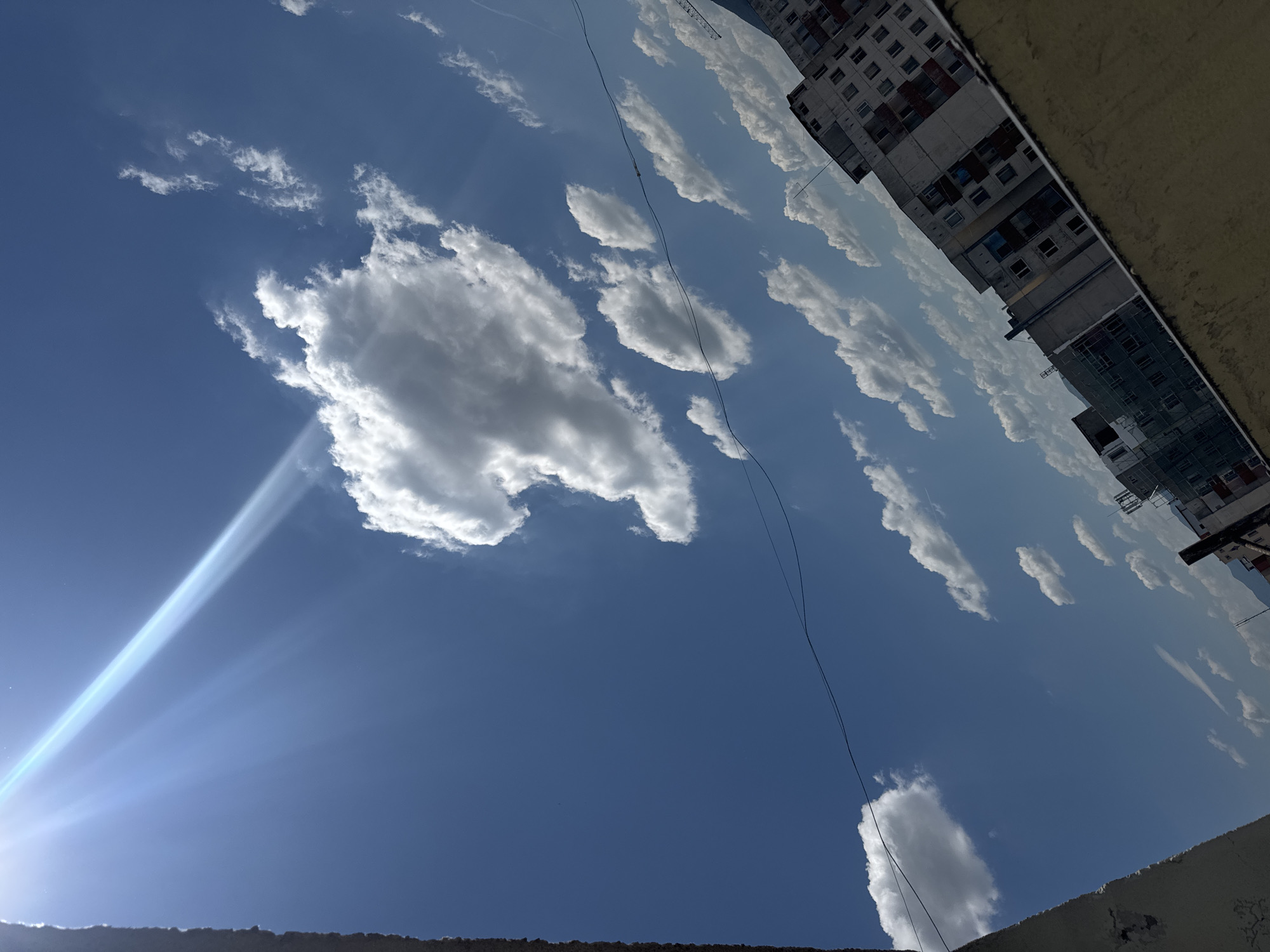 The image shows a bright, sunny sky with scattered cumulus clouds over Sofia, Bulgaria. The sun is visible in the lower left corner, emitting strong rays. High-rise buildings are partially visible in the upper right corner. A power line crosses the sky from top to bottom. The shot appears to be taken from ground level looking upwards.