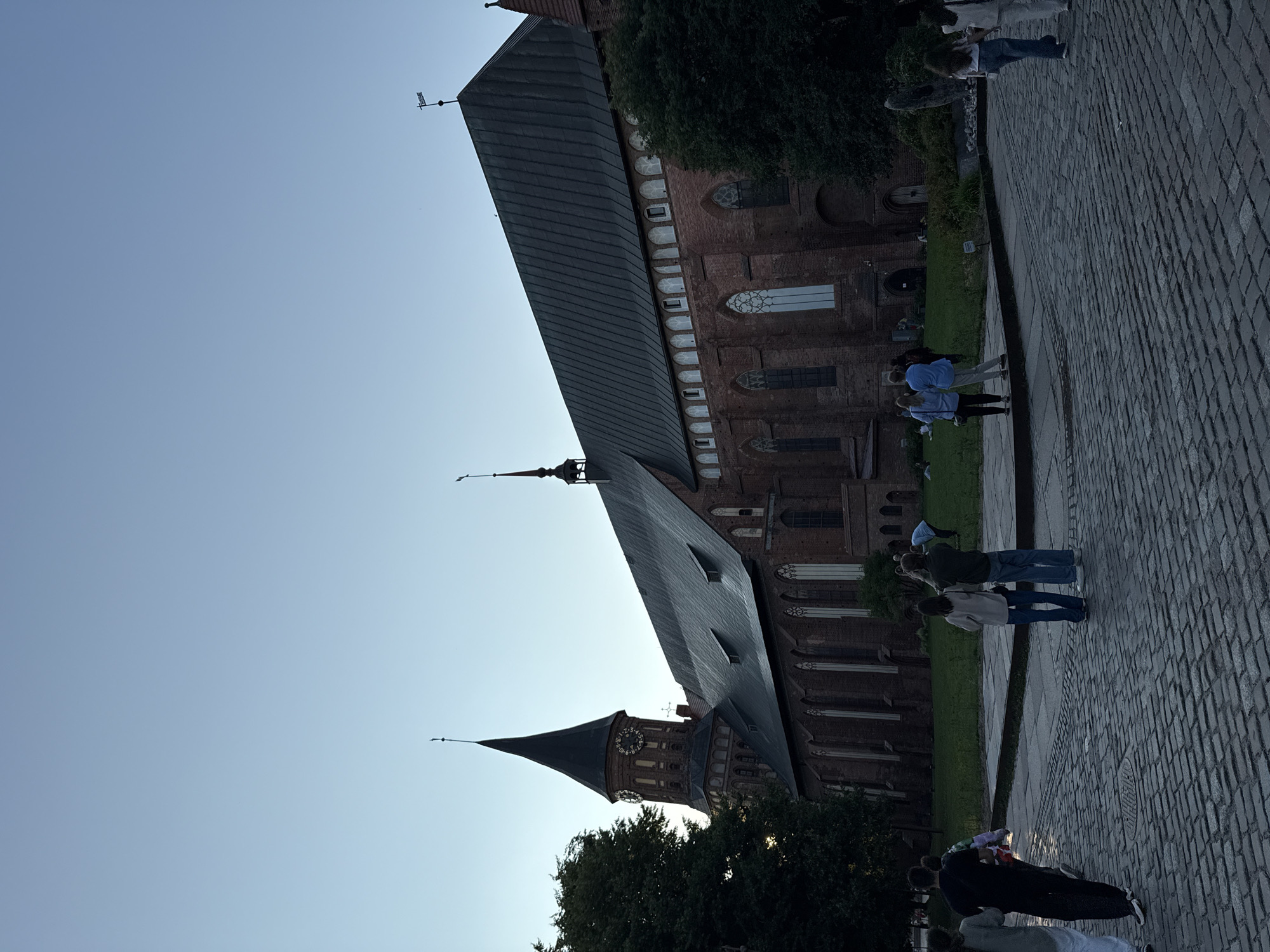 A low-angle, full shot captures the Kaliningrad Cathedral in Kaliningrad, Russia, under a clear, pale blue sky. The cathedral, constructed of red brick, features a tall, dark spire with a clock face and is partially obscured by dark green trees on the left side of the frame. The building's roof is dark gray with visible dormer windows. In the foreground, a group of people are walking on a cobblestone plaza. Some are facing away from the camera, walking towards the left, while others stand in small groups, looking at the cathedral or interacting with each other. The overall scene suggests a late afternoon or early evening setting with subdued lighting.