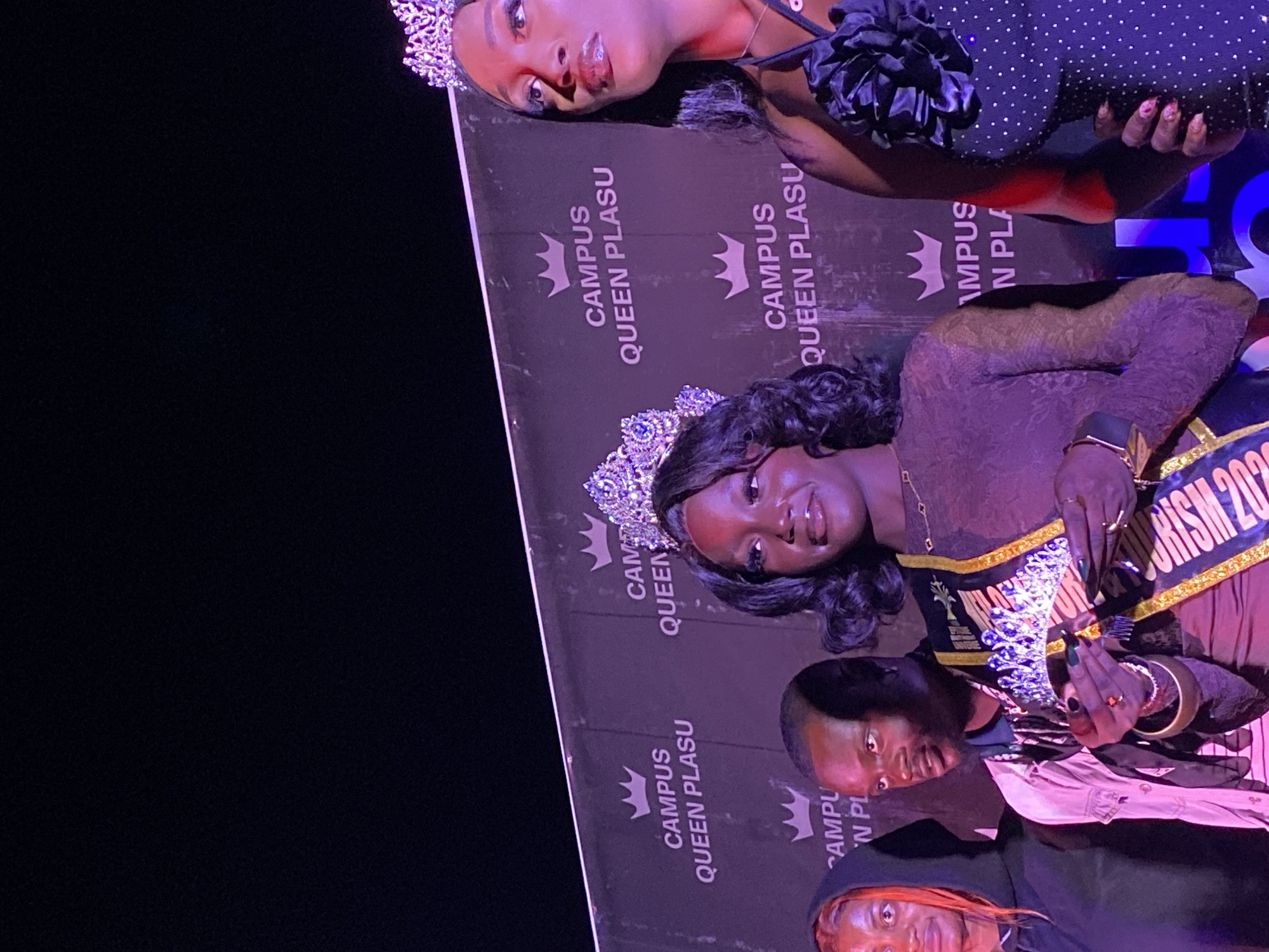 The image shows a group of people posing in front of a "Campus Queen PLASU" banner. Two of the women are wearing crowns, and one is wearing a sash that says "Tourism 202". A man is visible in the bottom center of the image. There is a dark background on the left side of the image. The location in the city of Durimi, Nigeria is not possible to determine from the context.