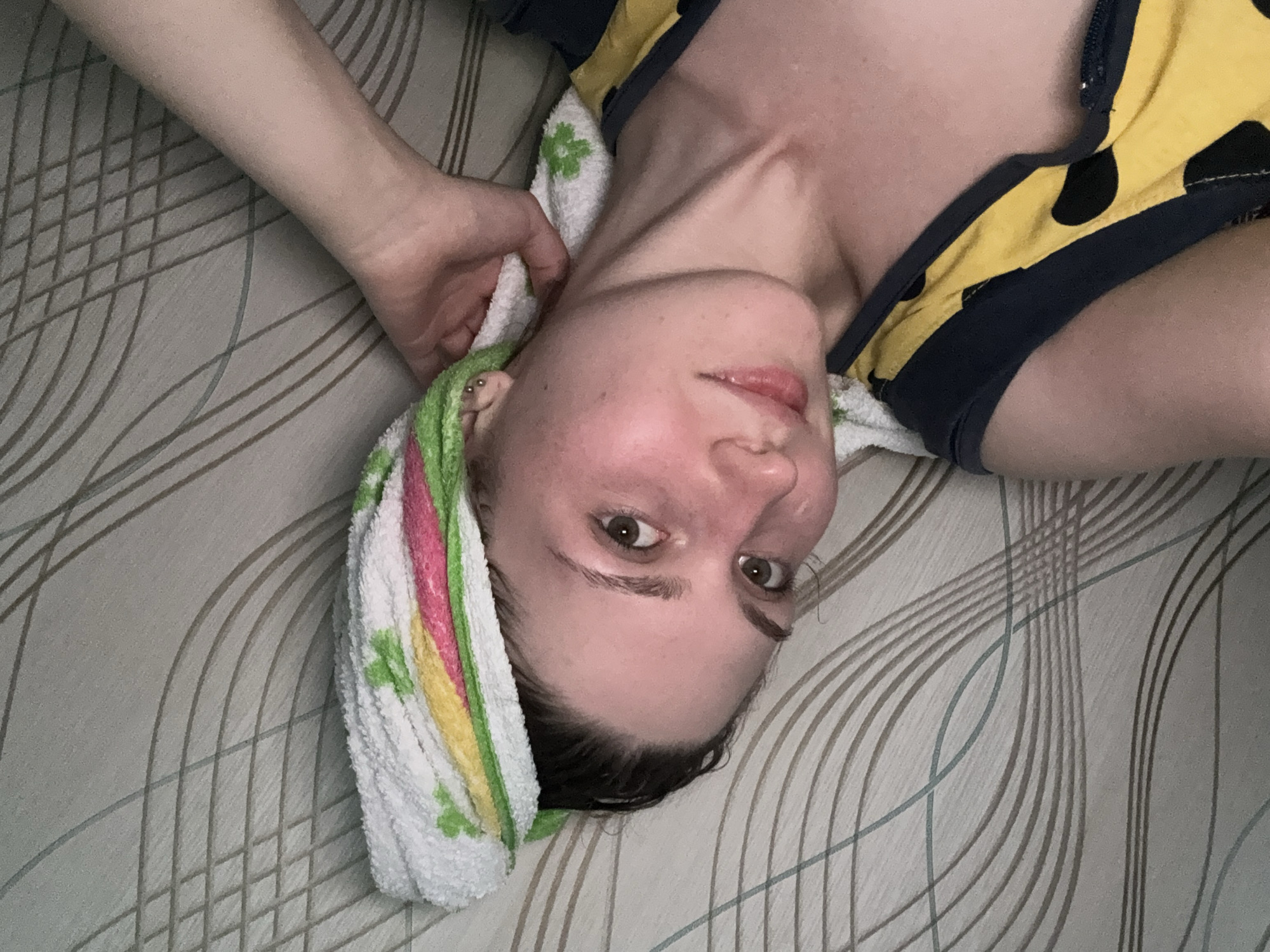 In an indoor scene in Дресвяново, Russia, a young woman is shown lying on her side, facing the camera. She has dark hair, styled with a towel wrapped around her head, featuring green, yellow, and pink stripes and a floral pattern. Her skin appears flushed, and she is wearing a yellow and black spotted top. Her left arm is visible, extended and resting on the towel. The background is a textured surface with a pattern of thin, curved lines in shades of gray and brown.