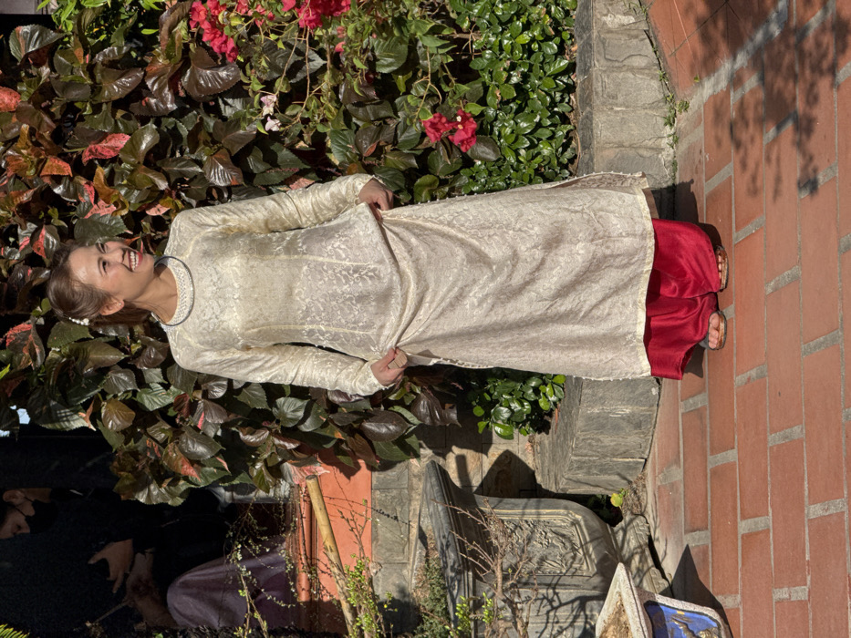 An adult female, appearing in her 30s, stands centrally on a terracotta-tiled patio, facing slightly right with a broad smile. Her dark hair is pulled back, adorned with a small white pearl-like clip on the right side. She wears a cream-colored, long-sleeved Ao Dai with an embossed floral pattern, featuring a high neck with white beading. Beneath the Ao Dai, she wears loose, vibrant red silk trousers and brown sandals. Her hands lightly hold the lower sides of the Ao Dai tunic.
In the shaded upper-left background, two additional individuals are partially visible: one is seated, wearing dark clothing and a black face mask; another figure wears lighter, possibly purple, attire.
The background consists of dense foliage, including broad-leafed plants with dark green and reddish-brown leaves, interspersed with red flowering plants, likely bougainvillea, on the right. A low stone retaining wall of grey, irregular stones supports the foliage. To the left, a multi-tiered structure of grey stone steps rises, with a red-orange tiled surface beyond. A large, ornate grey stone planter containing dry plant material sits on the patio in the lower-left, next to a smaller, blue-and-white patterned planter.
The scene is illuminated by bright sunlight, casting distinct shadows on the reddish-brown terracotta tiles of the patio. This is an outdoor garden or courtyard setting within a traditional Vietnamese architectural complex, located in the city of Nha Trang, Vietnam.