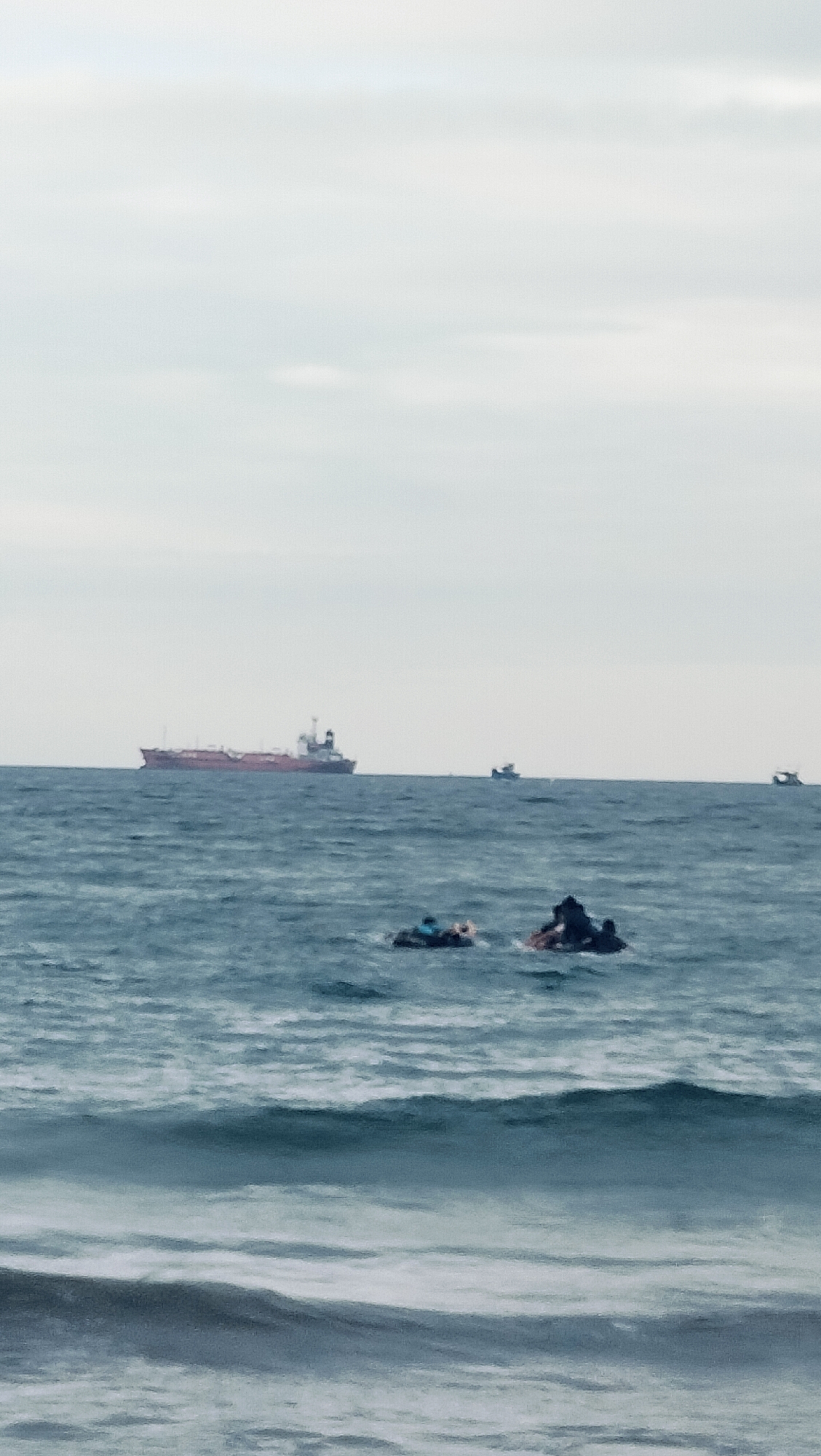 This image captures an overcast seascape in Lhokseumawe, Indonesia. The sky is a uniform light grey, indicating overcast conditions. The sea is a choppy, dark blue-grey with visible waves. In the immediate foreground, two dark, indistinct objects are floating on the water. The object on the left appears to be an inflatable ring or small raft, containing one or more individuals, one of whom shows a visible blue element. The object on the right is similar, also containing dark figures. Further out on the horizon, a large vessel, likely a tanker or cargo ship, with a reddish hull and a dark superstructure, is prominently visible. To the right of the large ship, two smaller boats are also present in the distance.