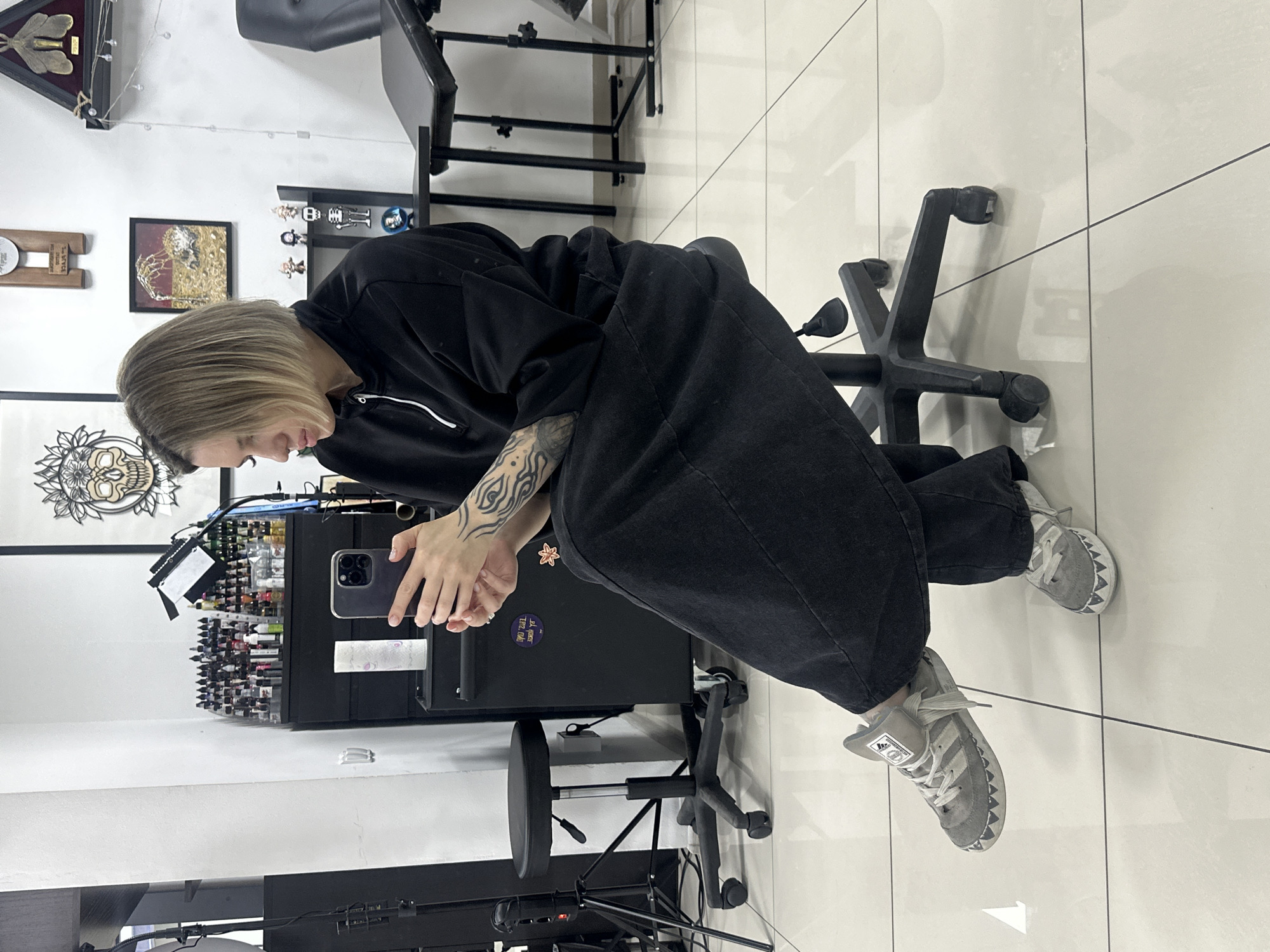 A female subject, approximately 20-30 years old, with short, light brown hair, sits on a black rolling stool with five caster wheels, positioned on a glossy white tiled floor in what appears to be a tattoo studio in Rostov-on-Don, Russia. She is dressed in a loose-fitting black long-sleeved top with a quarter-zip collar and wide-legged black pants. Her right forearm displays extensive tribal-style tattoos. She holds a dark-colored smartphone, possibly an iPhone, with a triple-lens camera array, in both hands, gazing downwards at its screen. Her feet are crossed at the ankles, revealing gray Adidas high-top sneakers with white laces and a distinctive white shark-tooth pattern along the front edge of the sole.
In the background, a white wall transitions into a black section on the left. To the left of the subject, a black stand with a ring light is partially visible. Behind her, a black cabinet or workstation is laden with numerous small bottles of tattoo ink in various colors arranged on tiered shelves. Another black stool with a round padded seat and caster wheels is visible to the left of the cabinet. Artworks adorn the white wall: a framed cutout of a skull with a floral wreath, a wooden plaque, a framed piece depicting a skeletal hand, and a triangular shadow box containing an insect specimen, possibly a moth. Below the skeletal hand artwork, small figurines are displayed on a shelf. To the right, a black medical-style table or tattoo bed with a headrest is positioned against the wall. Overhead, an LED panel light is mounted on a stand. The floor reflects overhead lighting and partial reflections of the room's fixtures.
