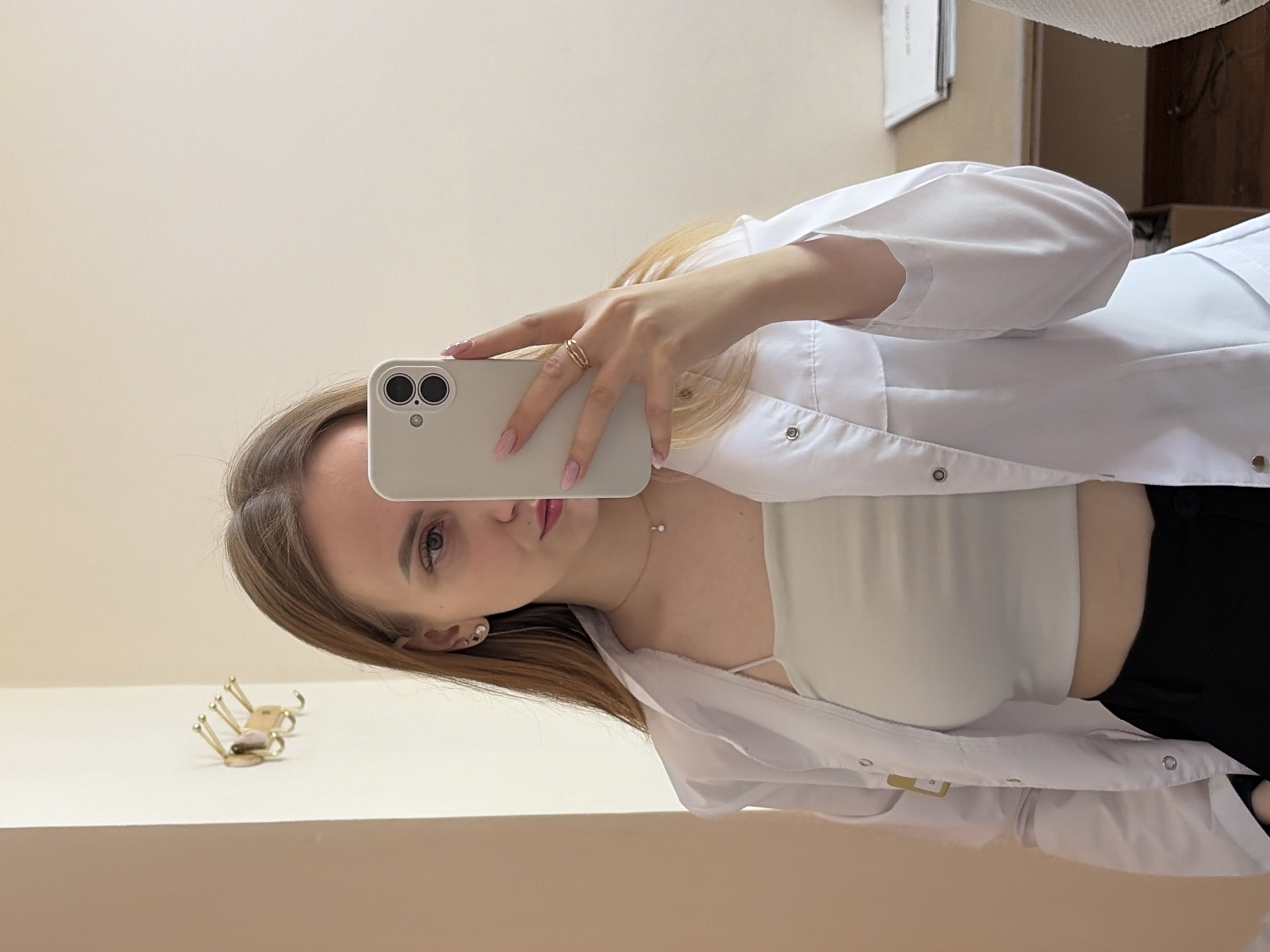 A young woman with light brown hair is taking a selfie in a mirror. She is wearing a white button-down lab coat over a light-colored top and black pants. Her face is partially obscured by a light-colored smartphone. She is wearing a gold ring and a necklace. Behind her, a wall and a cabinet are visible. On a surface reflected in the mirror is a small gold-colored object. The picture was likely taken indoors. There is no information in the image that confirms the location as the city of Chaikovsky, Russia.