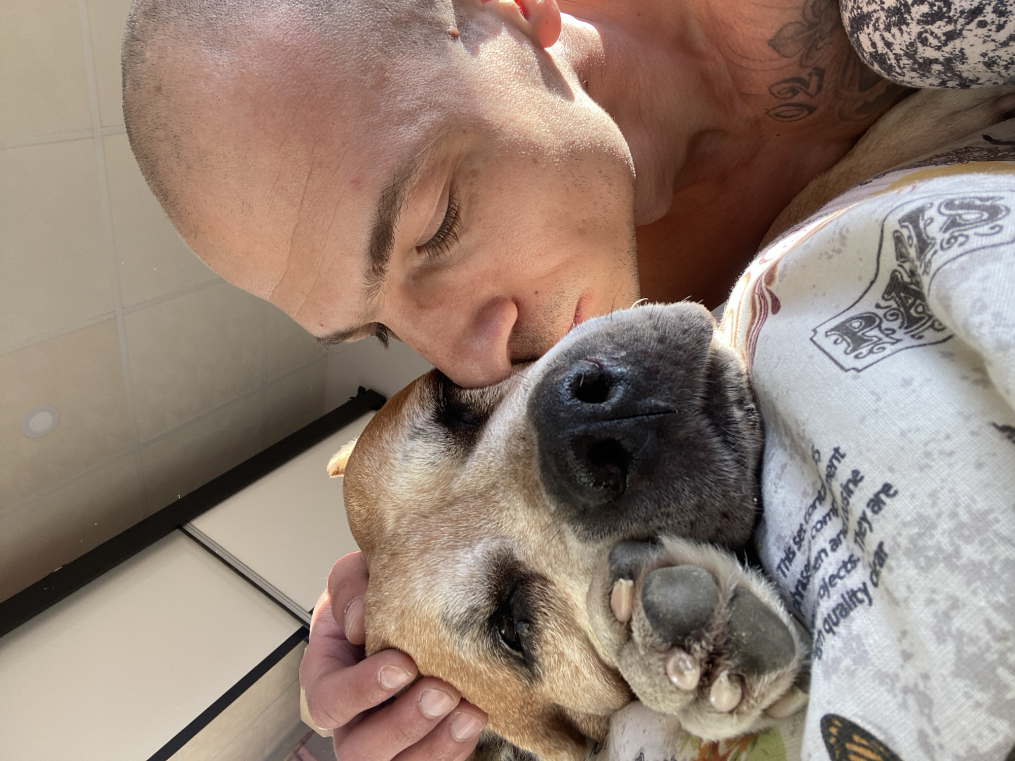A close-up shot shows a bald man with tattoos on his neck nuzzling a tan dog. The man’s eyes are closed, and his nose is touching the dog’s head. The dog’s nose is black, and its paw is visible. The dog is covered in a printed blanket. The scene appears to be indoors, as a cabinet can be seen in the background. There are no discernible details to place the location in Pleven, Bulgaria.