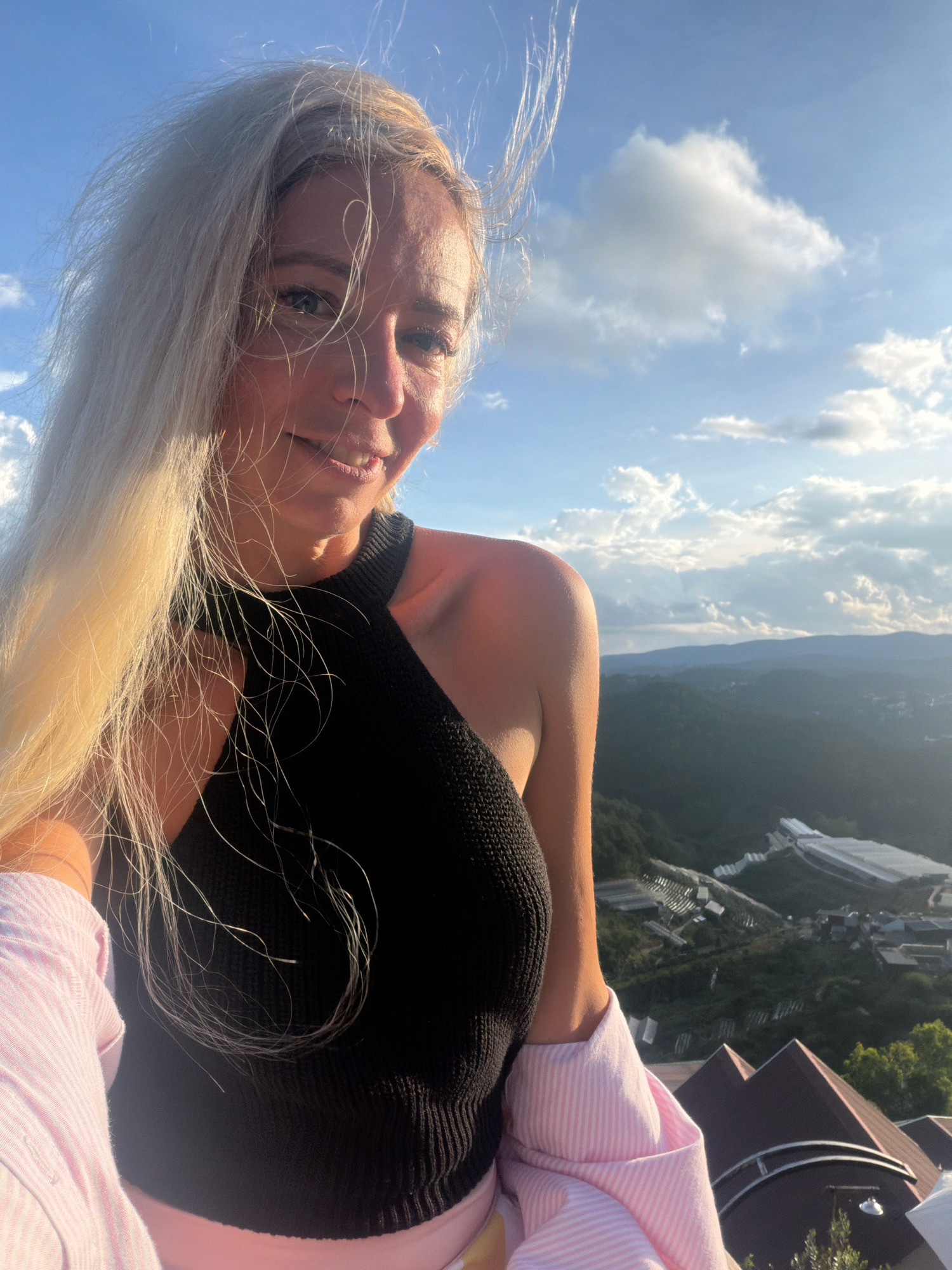 A blonde woman with long, blonde hair is taking a selfie outdoors, against a backdrop of a cloudy blue sky and a verdant, mountainous landscape. She is wearing a black halter top and a pink striped shirt. The landscape features rolling green hills dotted with buildings and what appear to be greenhouses or farms. The sun is low in the sky, casting a warm glow on the scene. The location is identified as Xuan Thanh, Vietnam.