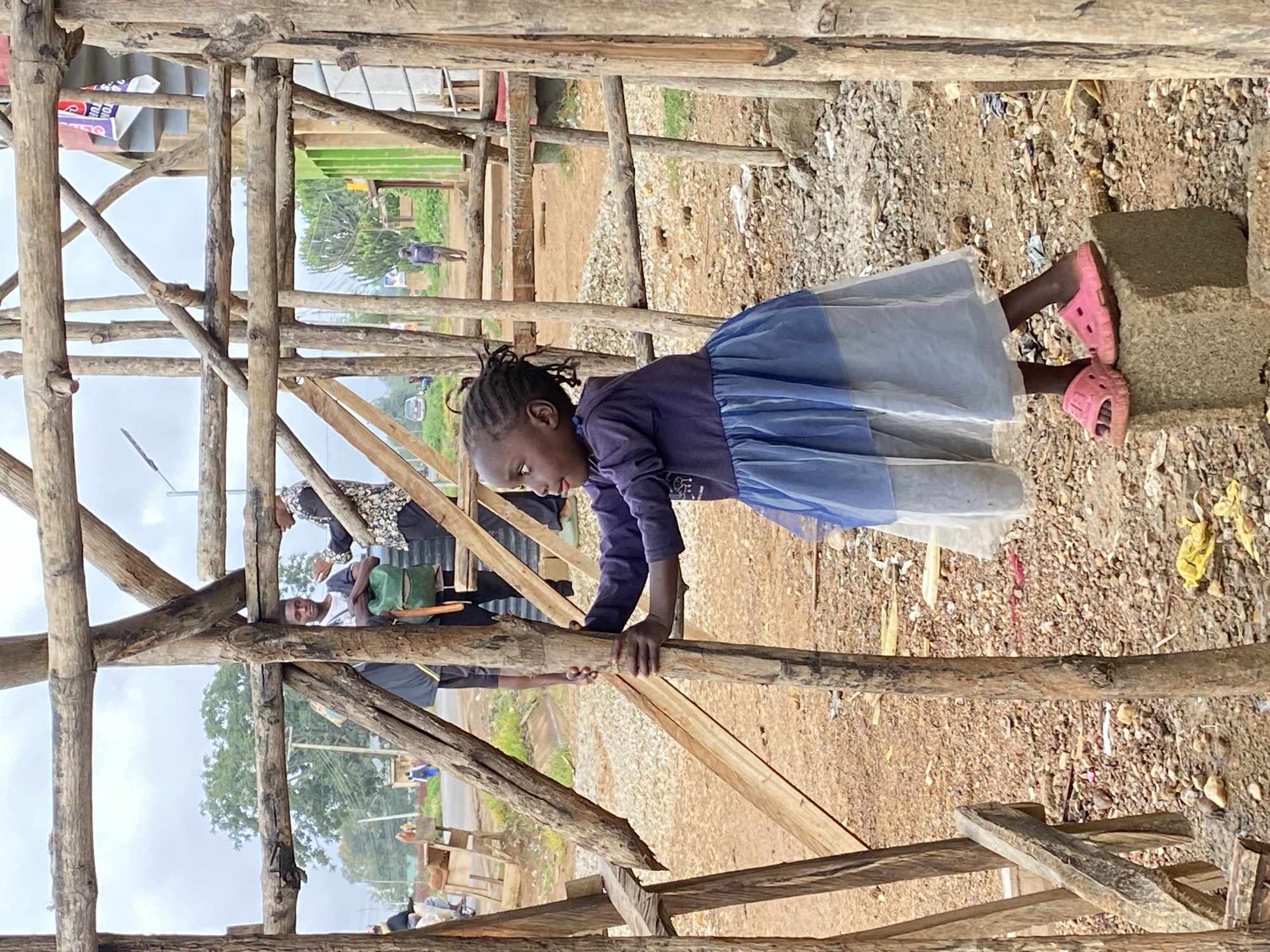 A young Black child in Riyom, Nigeria, is depicted interacting with a rustic wooden structure, likely a scaffold or temporary framework. The child, seen from the side, wears a long-sleeved purple top with a faint white design, a layered blue and white skirt, and distinctive bright pink Croc-style sandals. They are leaning forward, grasping a horizontal wooden beam with both hands, focused downwards or slightly to the left.
The wooden structure, made of unrefined logs and branches, dominates the foreground and midground. The ground is a mix of dirt, gravel, and small stones, with some debris. In the background, other individuals are present: a woman in a black and white patterned top stands near the structure, and further back, a man in a white shirt and dark clothing is visible, accompanied by a green bag or backpack. Piles of gravel or construction materials are scattered on the ground.
The scene takes place outdoors during daytime, under an overcast sky, with soft, diffused lighting. Trees and what appears to be a road or path are visible in the distance. Although there are signs in the far background, any text or graphics on them are illegible. The image captures a moment of engagement within an active, possibly semi-urban or rural setting.