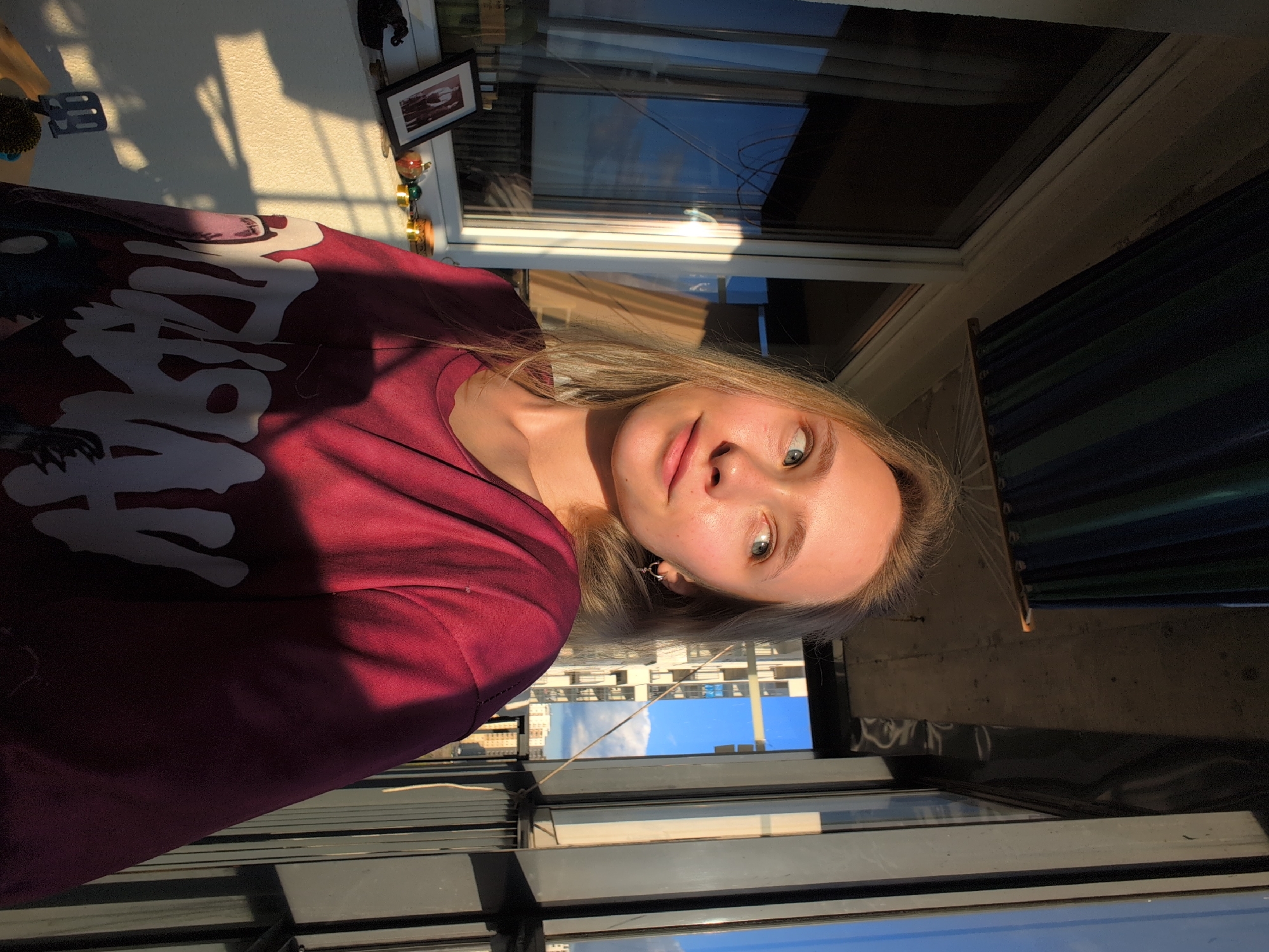 The image shows a young woman with blonde hair and blue eyes posing on a balcony in Bugry, Russia. The woman is wearing a maroon sweatshirt with a white graphic print. Sunlight is streaming in from the right, casting a shadow on her face and chest. In the background, there is a window with curtains, a shelf with various objects, and buildings visible through the balcony railing. The scene appears to be a casual, outdoor setting on a sunny day.