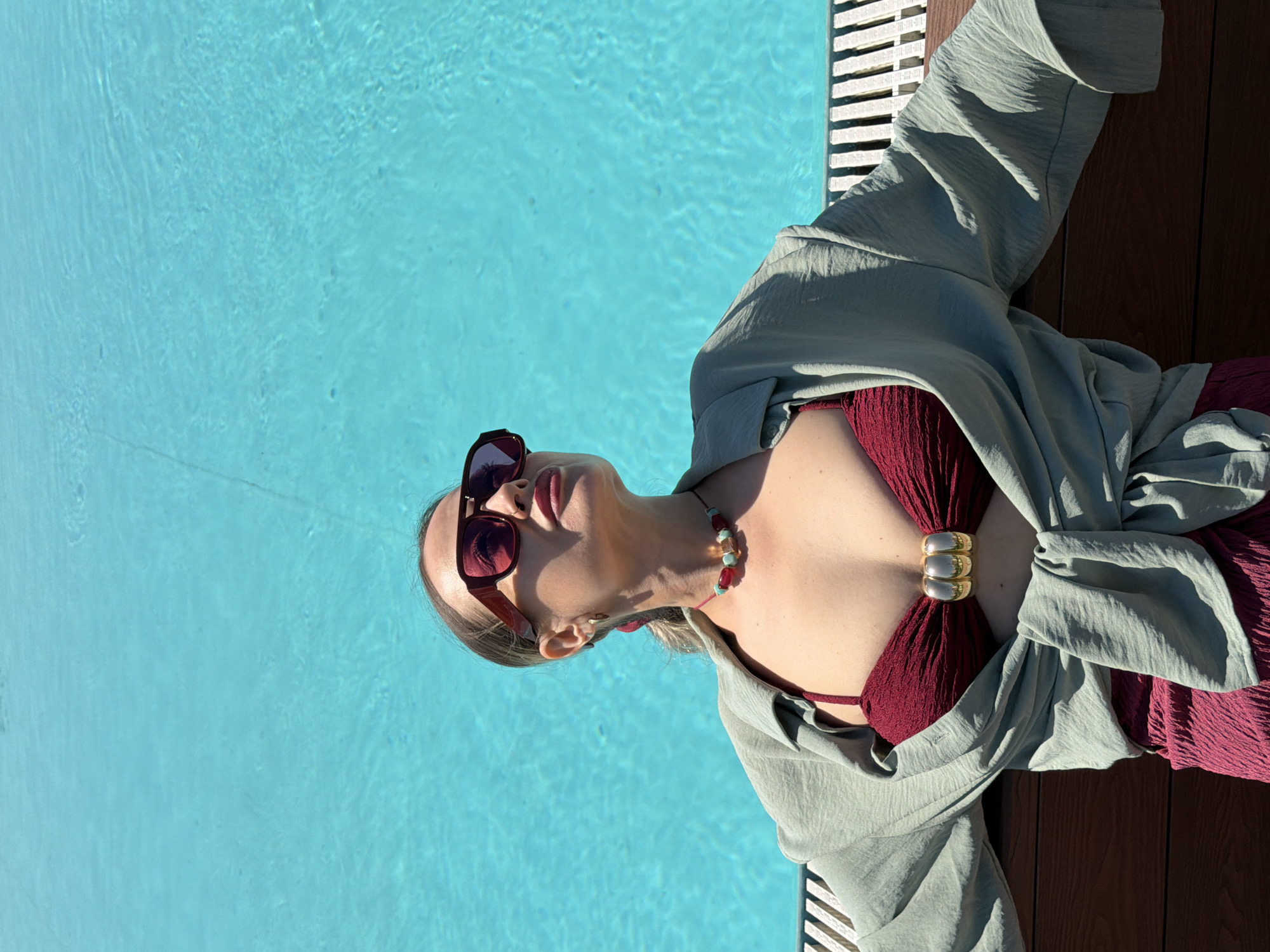 This high-angle, close-up image portrays a woman relaxing poolside in Krasnoyarsk, Russia, under bright daylight. The woman, with blonde hair, is visible from the chest up, lying on her back with her head tilted slightly upwards. She wears dark red, rectangular sunglasses and pink lipstick. A necklace with red, green, and golden-amber beads adorns her neck.
She is dressed in a dark red, textured bikini top, which features a prominent gold, ribbed, three-ring-like embellishment at the center of the bust. A light sage green or pale olive-colored oversized shirt or cover-up is draped loosely around her upper body, suggesting a casual, relaxed style. Parts of a dark red textured garment, possibly a skirt or sarong, are also visible.
The background is dominated by a clear, vibrant blue swimming pool, displaying gentle ripples and reflections across its surface. To the right of the woman, a dark brown wooden deck or poolside flooring, made of parallel planks, runs along the edge of the pool. The strong, direct sunlight creates sharp highlights and shadows, indicating a sunny day, likely mid-day or afternoon, contributing to a tranquil and leisurely atmosphere. There is no visible text in the image.