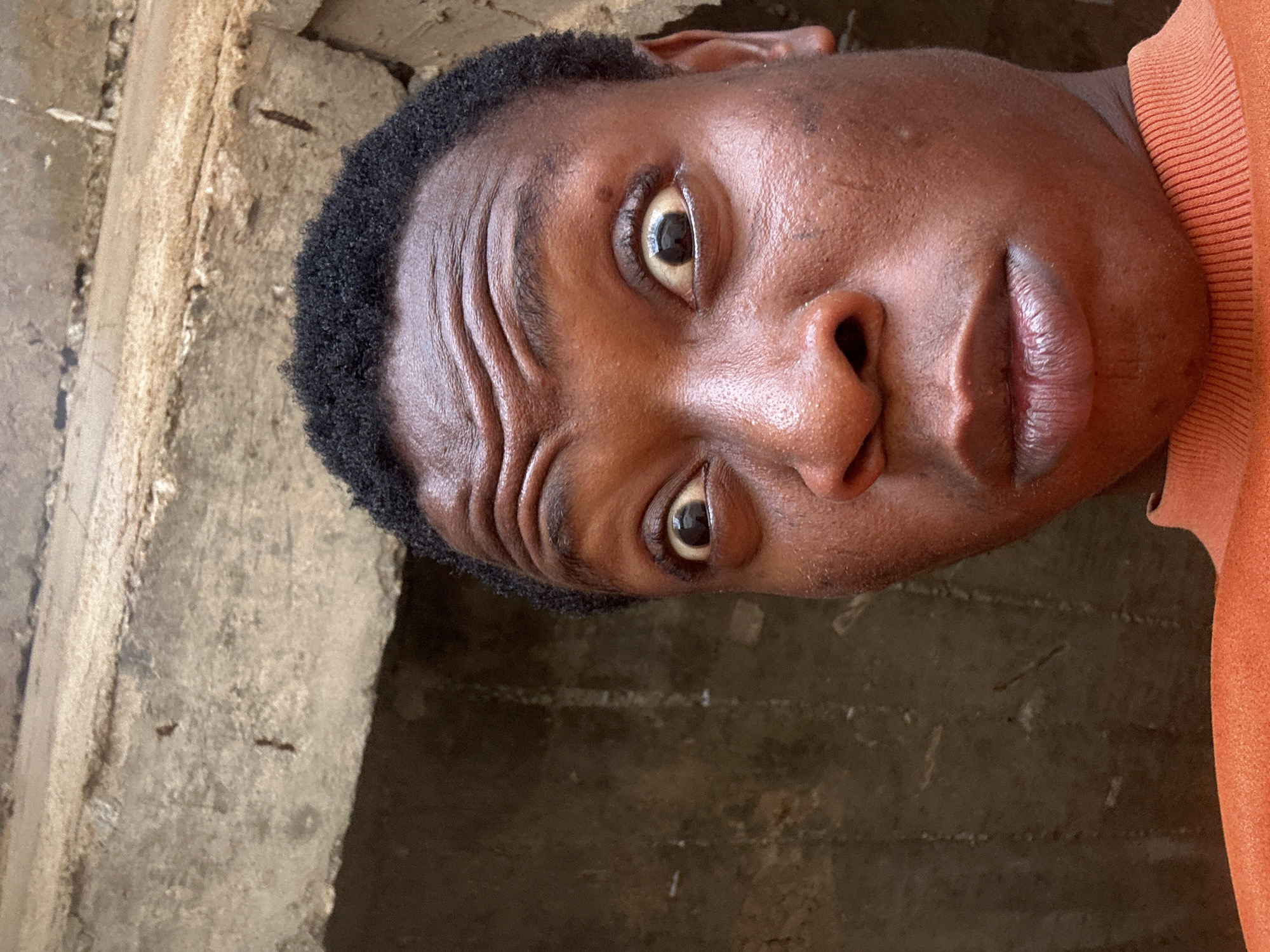 A medium close-up shows a Black man from a low angle, his face tilted and appearing to look upwards. The man has short, dark hair and dark eyes that seem wide open. His skin has a slightly wet sheen. He wears an orange garment near the edge of the frame. The background features a textured, gray and brown wall, possibly concrete or masonry, appearing aged and weathered. The location is Unknown, Unknown.