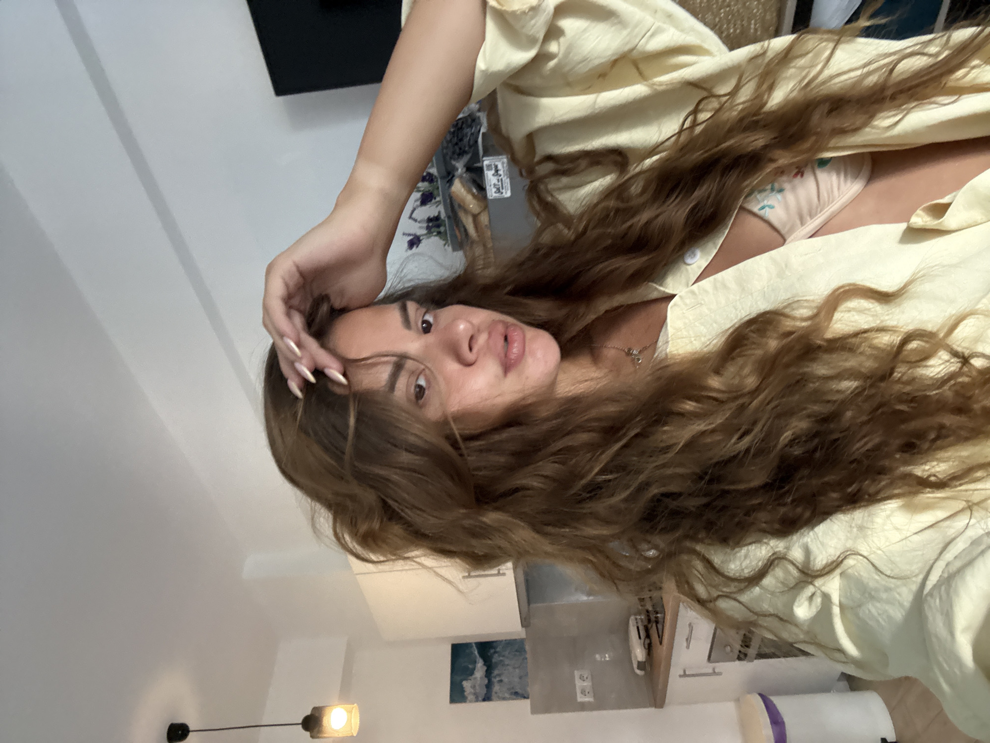A woman with long, wavy brown hair is shown in a close-up shot, with her right hand touching her head. She is wearing a light yellow open shirt over a patterned bra. The background appears to be a kitchen area with white cabinets and a countertop. A light fixture hangs from the ceiling on the left side of the image. The scene is indoors.