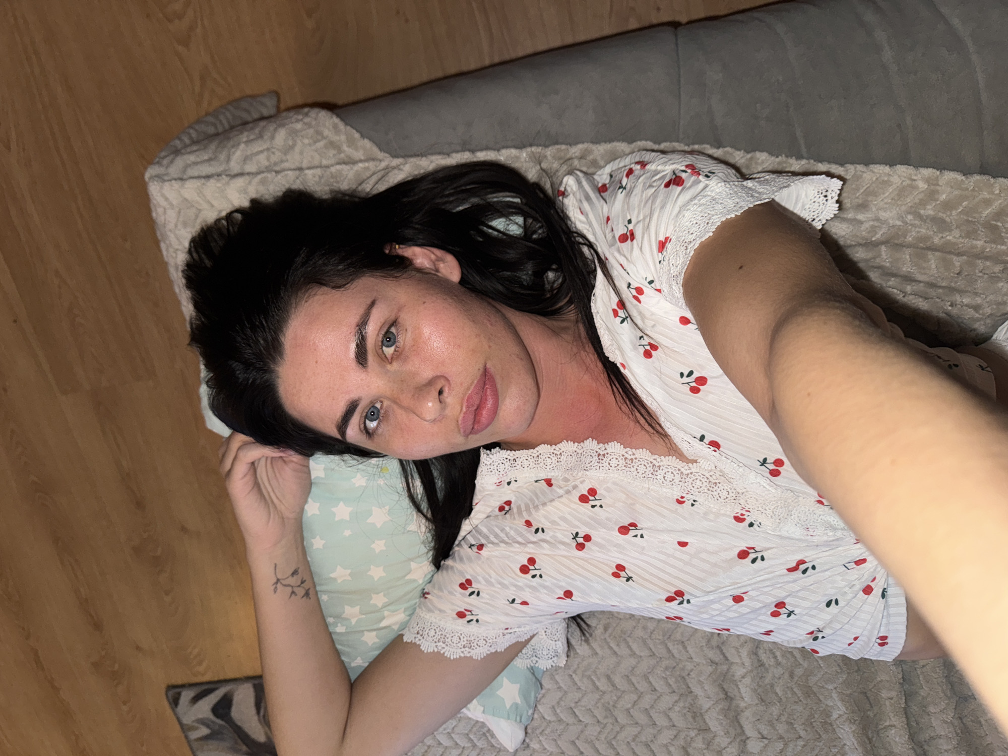 An image captures a woman lying down indoors, looking directly at the camera. She has long, dark hair, blue eyes, and full lips. She is wearing a white top adorned with red cherry patterns and white lace trim on the sleeves and neckline. A small, delicate tattoo resembling a sprig of leaves or flowers is visible on her left wrist.
She is resting her head on a light blue pillow decorated with white stars. The woman is situated on what appears to be a grey sofa or bed, covered by a textured, light-colored blanket. Part of a wooden floor is visible in the background, to the left of the woman. The lighting suggests an indoor setting, likely in the evening or at night, as artificial light illuminates the scene. The overall mood is casual and relaxed. There is no visible text in the image. This scene takes place in Chaikovsky, Russia.