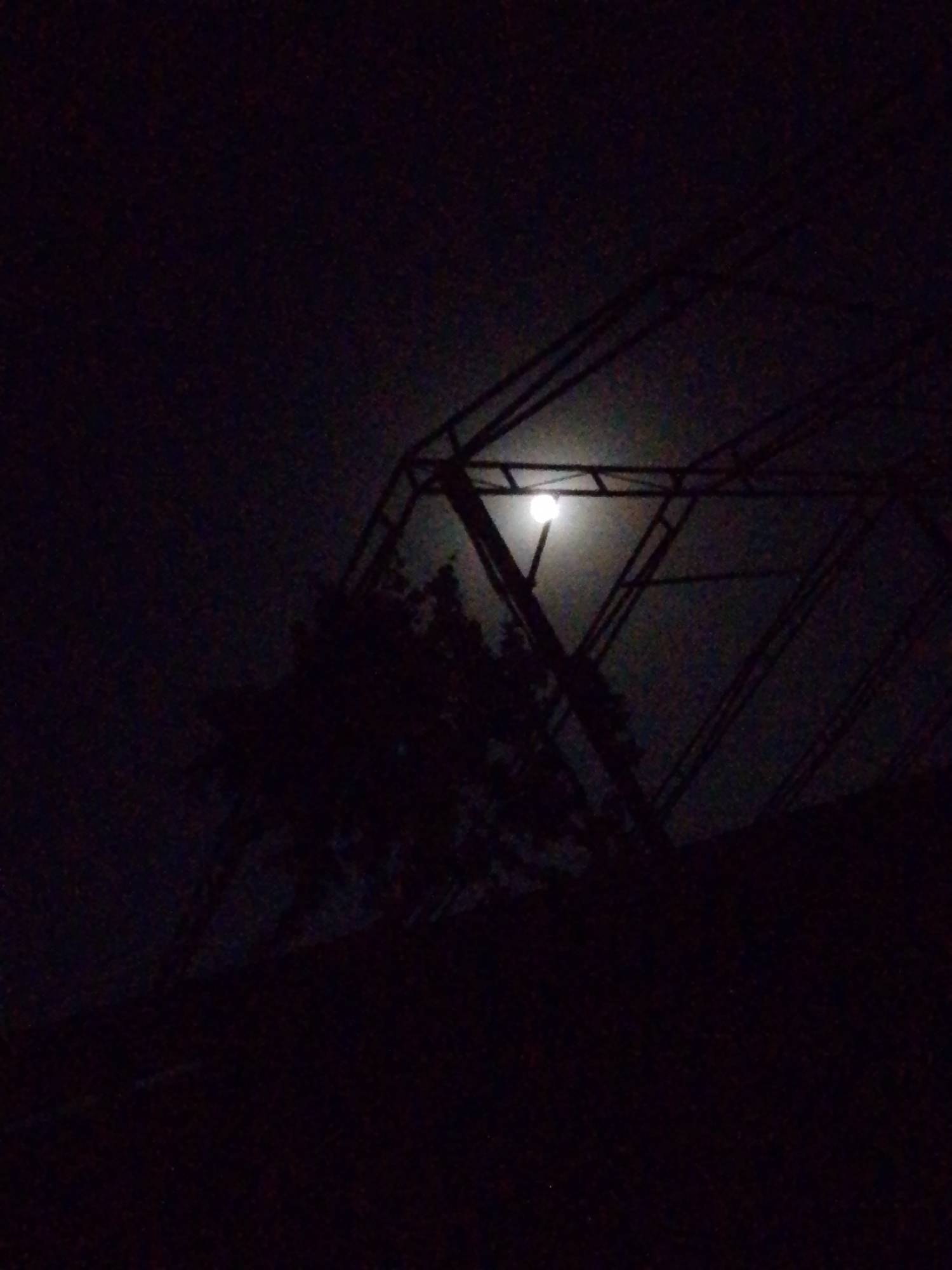 This is a nighttime photograph taken in Uchqizil, Uzbekistan. A bright full moon is visible in the dark sky, partially obscured by a large, geometric metal structure. The structure appears to be an angular framework, possibly part of a building or an art installation. Silhouetted against the moon and the dark sky are the branches of a tree. The overall scene is dark and atmospheric, with the moonlight providing the primary illumination. The image has a grainy quality, typical of low-light photography. There are no people visible in the photograph. The environment appears to be outdoors, likely in an urban or semi-urban setting given the presence of the man-made structure.