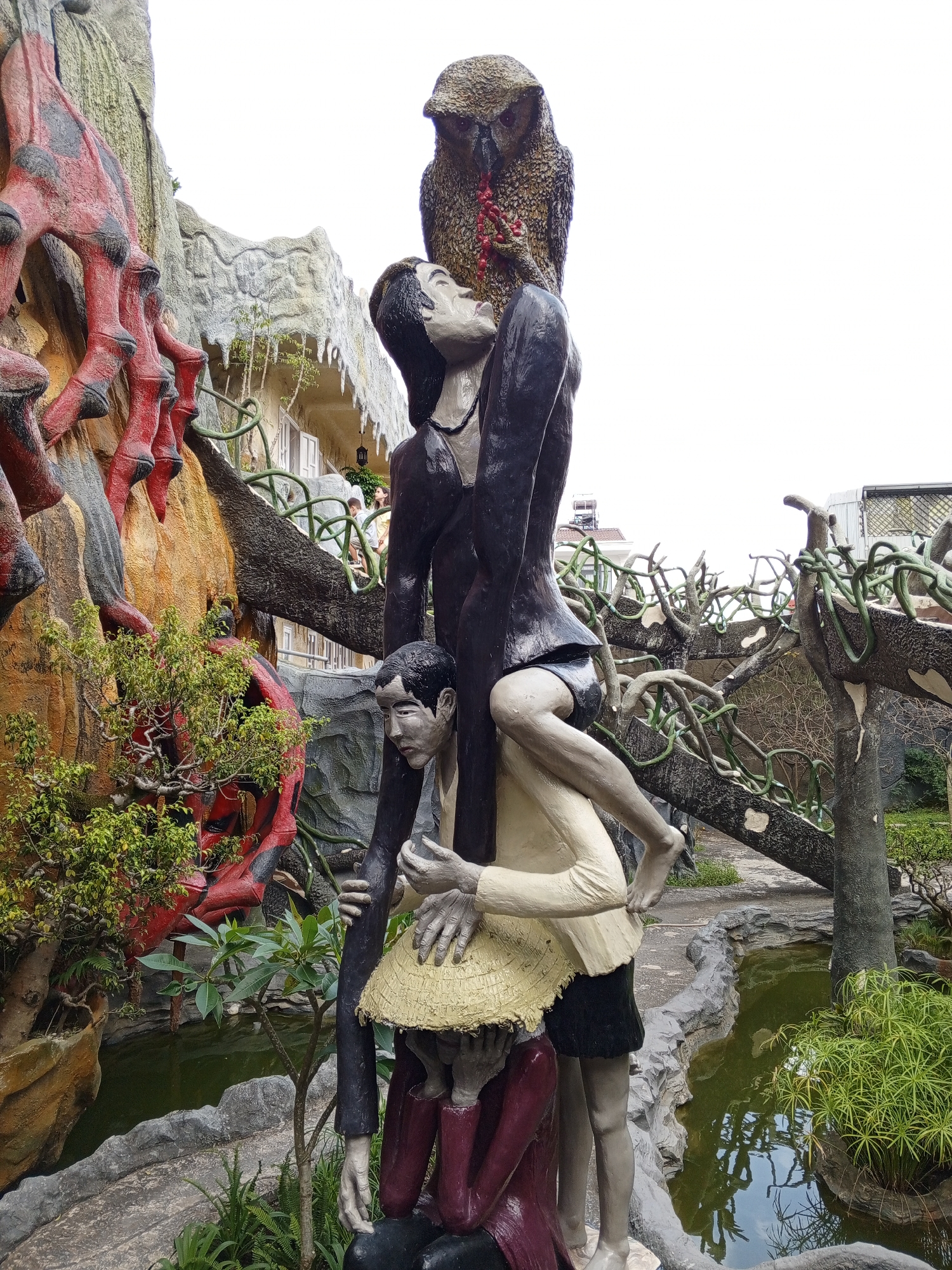The image shows a surreal sculpture in Da Lat, Vietnam, which appears to be located in a garden setting. The sculpture is a tall, tiered arrangement of figures. At the very top, an owl is perched, holding red berries in its beak. Below the owl is a figure of a person with a dark jacket and shorts, their face tilted upwards. This person is seated on the shoulders of another figure, who is wearing a cream-colored shirt and black shorts. The second figure's arms are around the person above them. Beneath these two figures, another person, wearing a maroon garment, is partly visible, suggesting they are at the base of the sculpture. The surrounding environment includes a pond with green water, rocky formations, and vegetation, including small trees and bushes. In the background, there are architectural elements and what appear to be other sculpted features, including a large red and black structure to the left. The overall impression is one of artistic expression with symbolic or allegorical elements, set within a visually dramatic landscape.