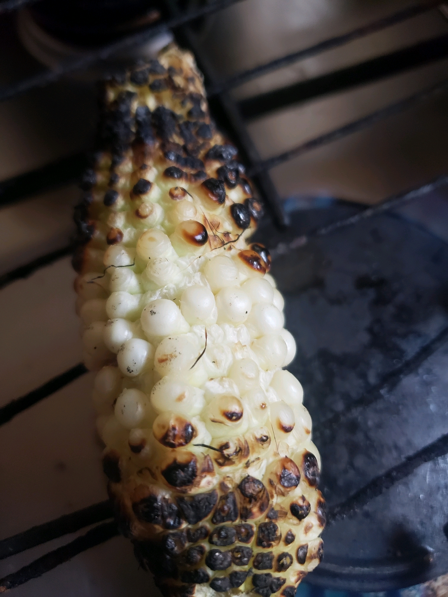 A close-up, eye-level shot shows a partially burnt cob of white corn, positioned vertically. The corn is situated on a dark, gridded surface, possibly a stovetop. The kernels are plump and white, with several showing signs of charring and browning, particularly at the tips. Some strands of corn silk are visible. The background is dark and out of focus, with hints of metallic elements suggesting kitchen appliances. The lighting is dim, casting soft shadows. The scene appears to be a casual, domestic setting, potentially in a kitchen in Sabon Tasha, Nigeria, where the corn is being prepared for consumption.