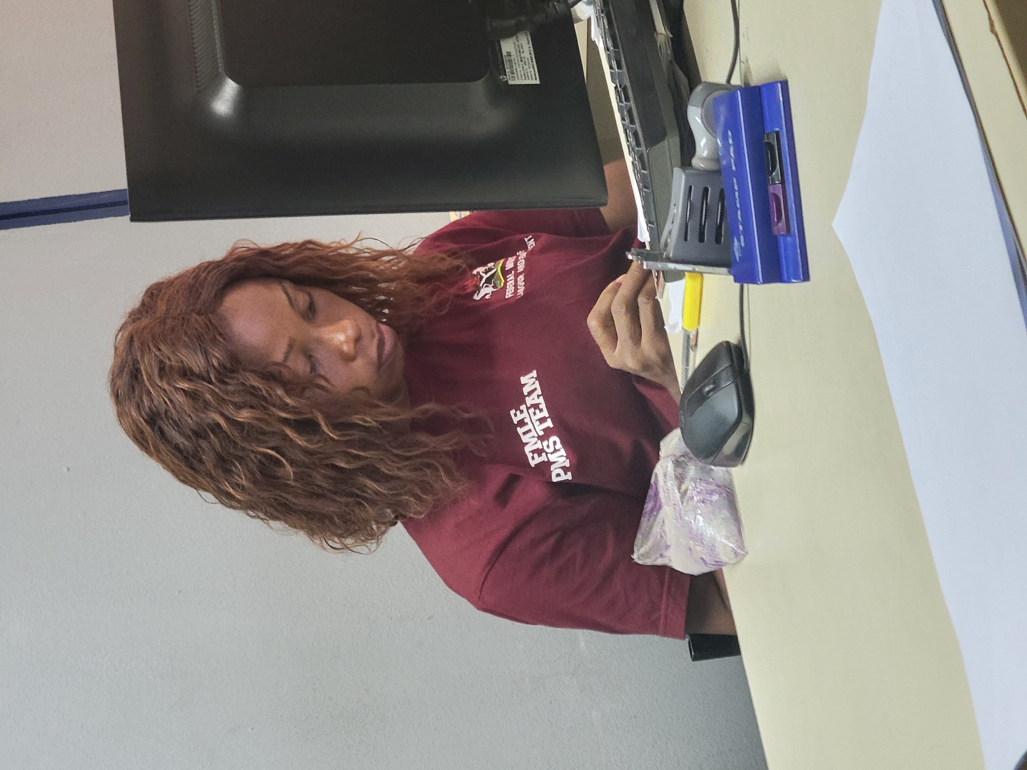 This image captures a woman with dark skin and long, wavy, reddish-brown hair, seemingly resting or sleeping at an office desk. She is wearing a maroon short-sleeved t-shirt. Her head is tilted to her right, and her eyes appear closed or mostly closed.
The setting is an indoor workspace, likely an office. A black computer monitor is positioned to her right, partially visible. On the light-colored desk to her right, various items are present, including a blue and grey stapler and a black computer mouse. A small, clear plastic bag, possibly containing light purple items, sits near the mouse. A white sheet of paper is also visible on the desk further to the right. Her right hand is near the base of the monitor, and a yellow object, possibly a screwdriver or pen, is partially visible near her fingers.
The background consists of a plain light grey wall, with a horizontal blue strip visible in the upper left portion of the image.
Visible text on the woman's t-shirt includes "FEDERAL AIRPOR..." (likely "FEDERAL AIRPORTS"), "LAGOS NIGERIA" (partially obscured), and "FMLE PMS TEAM," all in white lettering. This text, combined with the location context of Abuja, Nigeria, suggests the setting is an office within a Nigerian organization, possibly related to airport management. The woman's posture and closed eyes indicate she is either taking a brief rest or is deeply focused with her eyes closed during her workday.