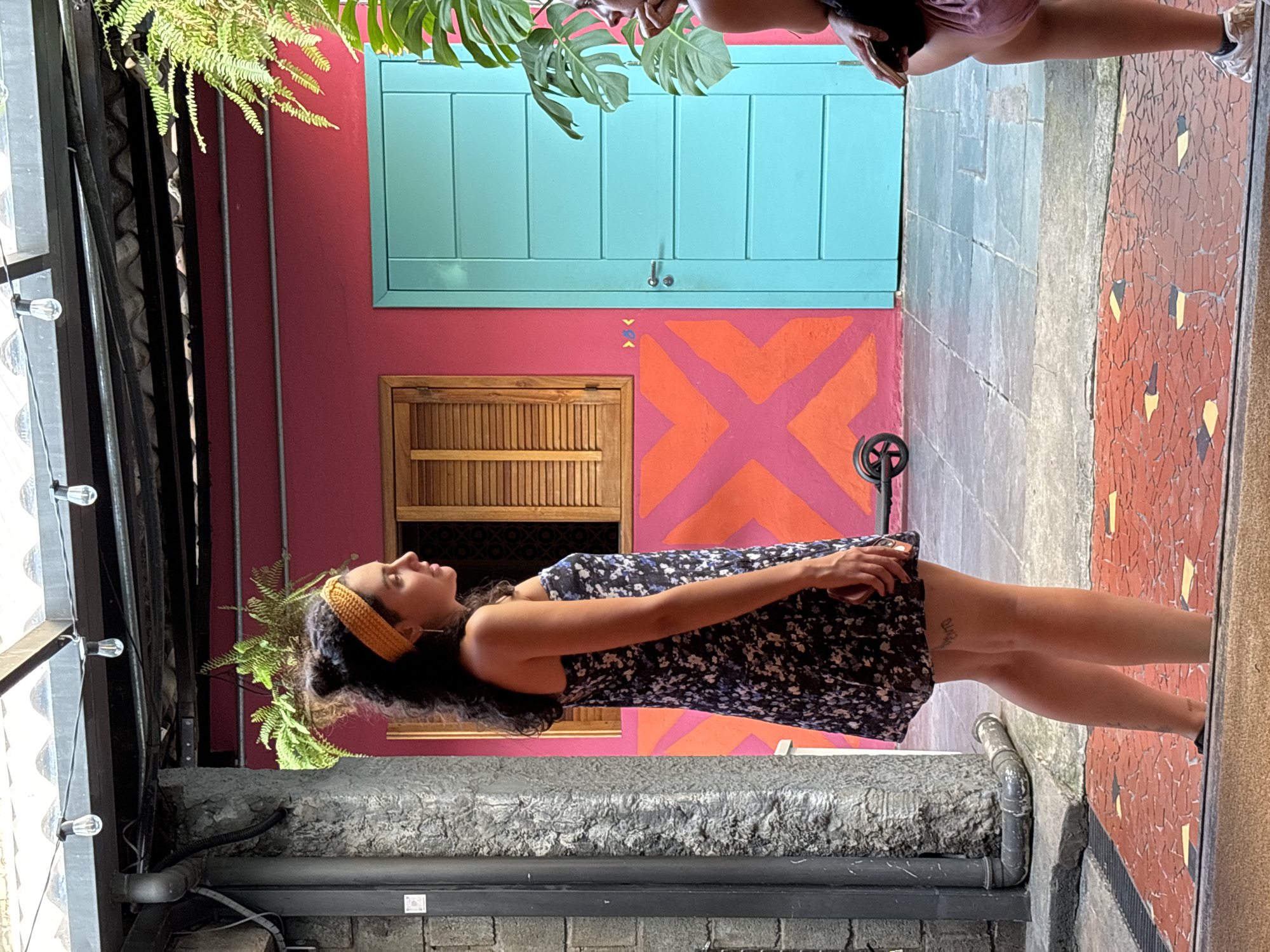 A young woman stands in a colorful courtyard. She wears a dark, floral-patterned dress and a yellow headband. Her dark hair is shoulder-length. She stands on a stone-tiled floor, next to a gray ledge with pipes and a light fixture above her. The wall behind her is pink with an orange "X" design. Above that is a blue door, and above the door is part of a person's body, possibly taking the photo. A fern hangs above her and on the left. The building is identified as in São Paulo, Brazil.