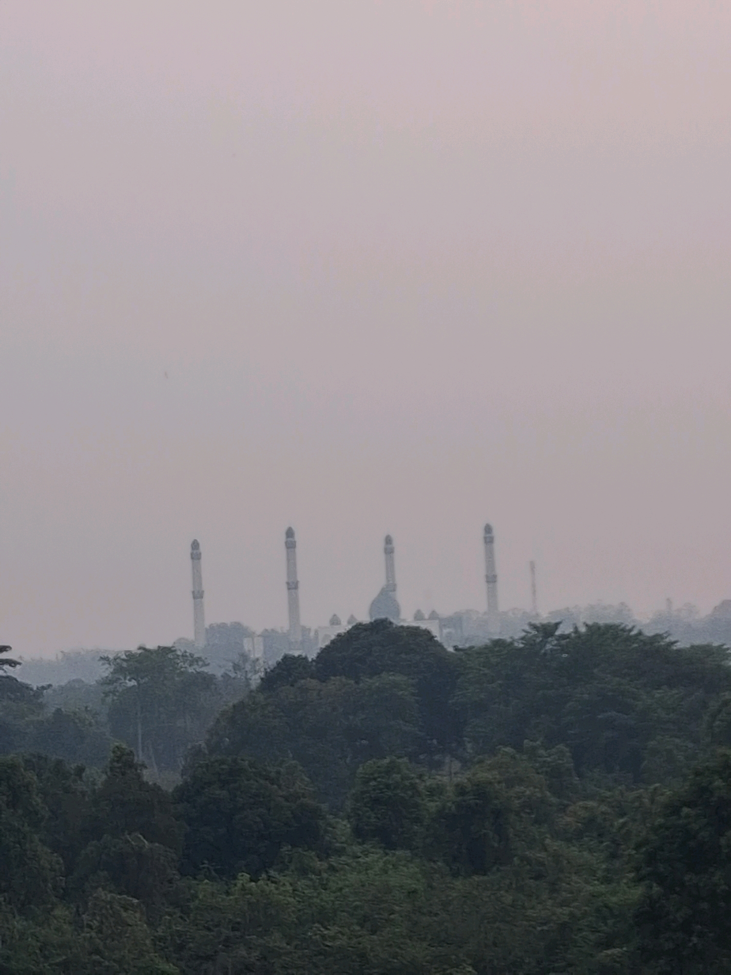 The image shows a hazy, overcast sky over a densely forested landscape. In the distance, several tall, slender minarets of a mosque are visible, rising above the trees. The minarets are light-colored and appear to be made of stone. The surrounding trees are a mix of dark green and lighter green, with some appearing to be taller than others. The overall impression is one of a serene and somewhat mystical scene, possibly captured during sunrise or sunset. The location is identified as Pangkalan Kerinci, Indonesia.