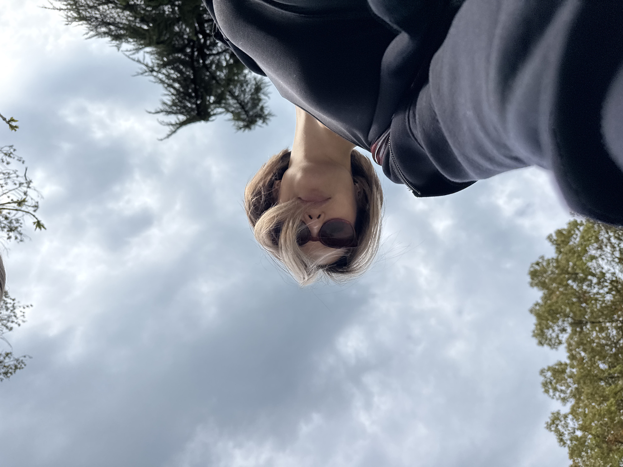 A person with blonde hair and sunglasses stands with their head facing down towards the camera. The person is wearing a black hoodie. The background consists of a cloudy sky and trees. The location is in the city of Unknown, Unknown.